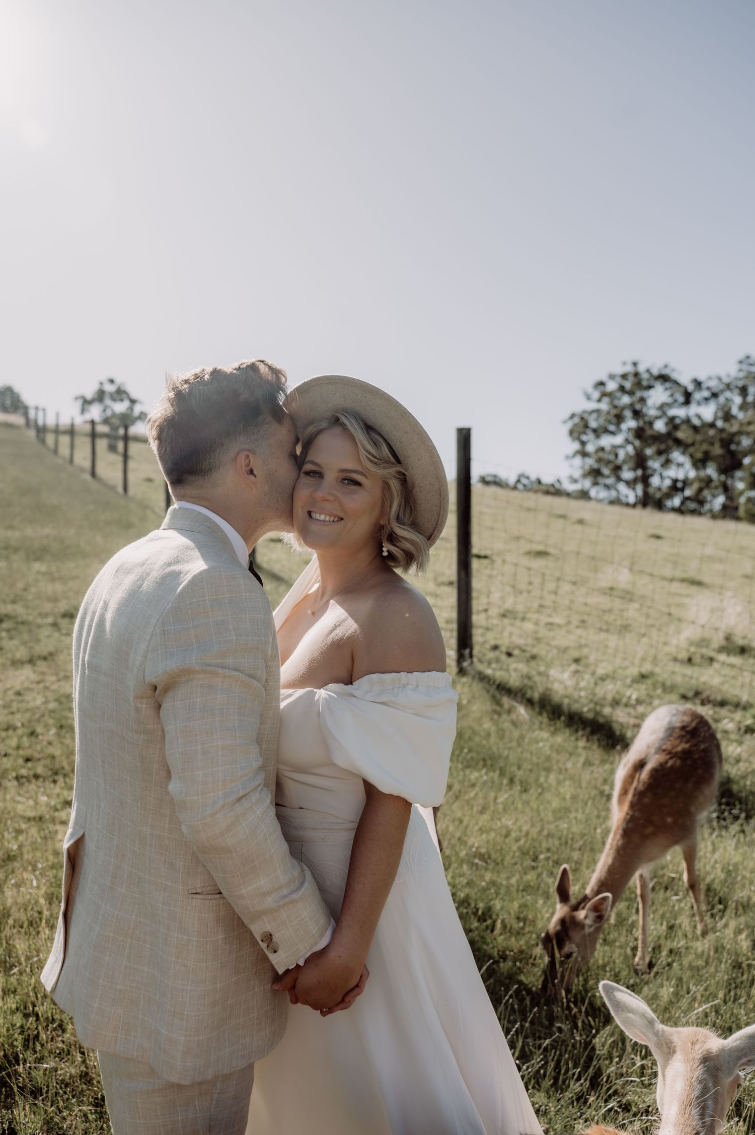 A couple shares romantic moments in a field while curious deer graze in the background.
