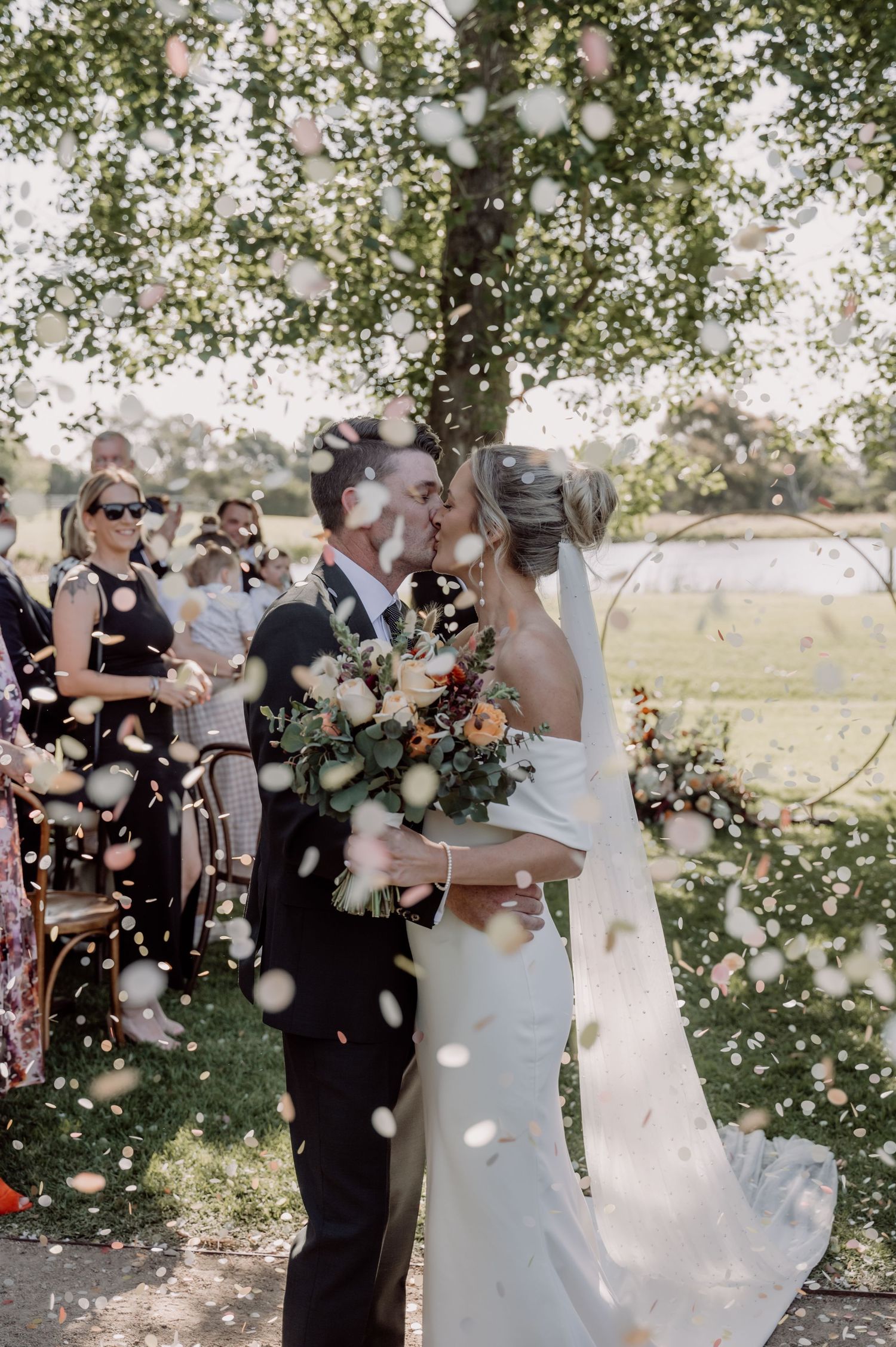 Newlyweds share a romantic kiss under falling confetti at an outdoor wedding ceremony as guests look on.