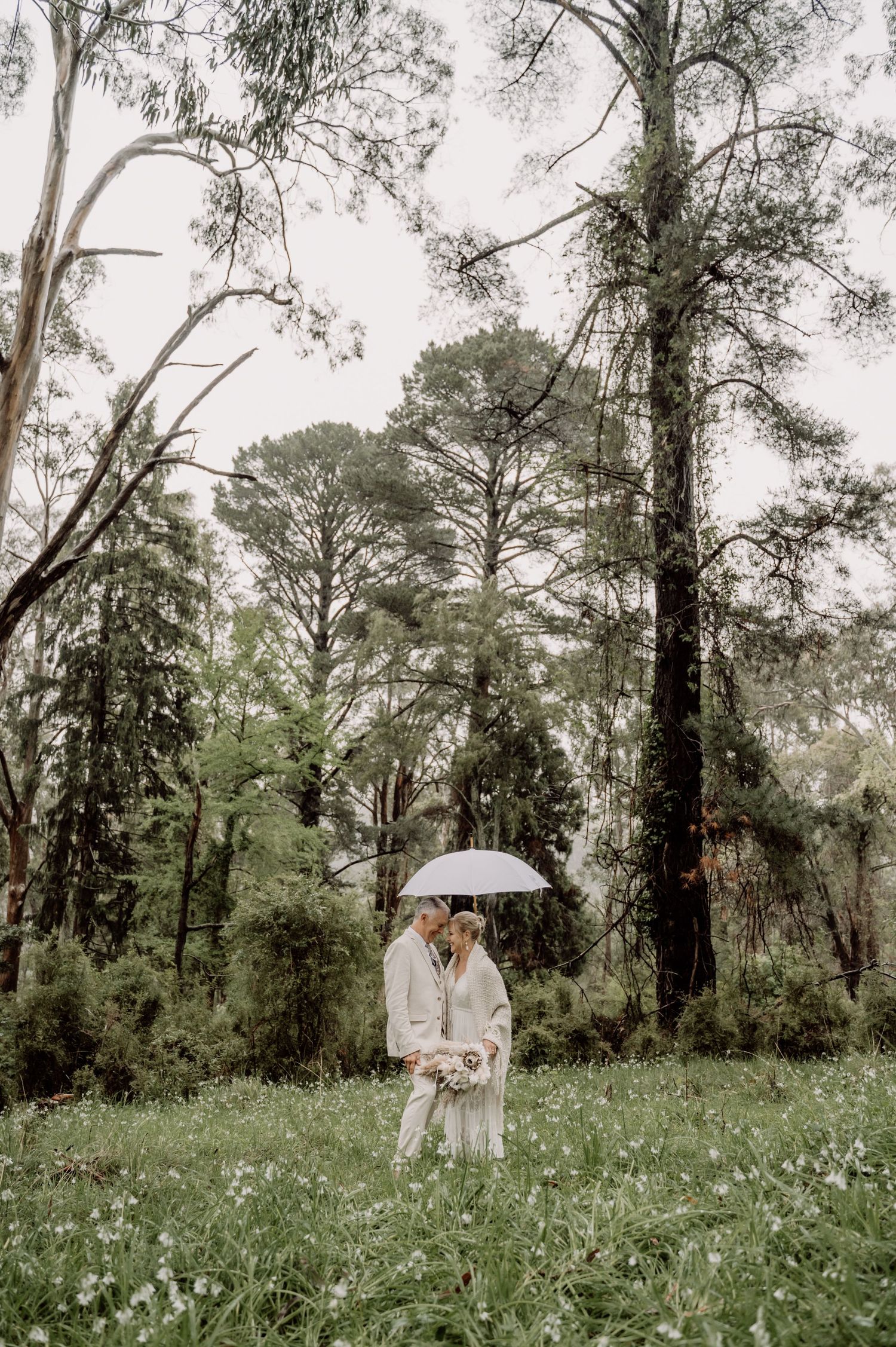 A couple sharing a romantic moment under a white umbrella in a lush green forest setting during an overcast day.