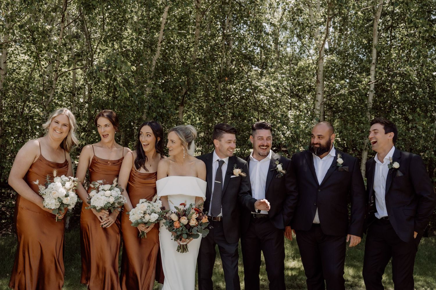 Wedding party in formal attire stands together outdoors among birch trees holding bouquets of white flowers.