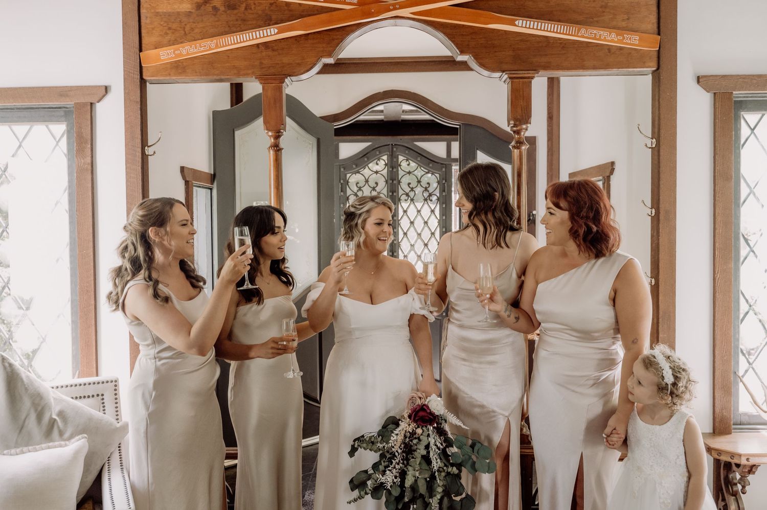 A bridal party in light colored gowns gathers by a window in a rustic indoor setting.