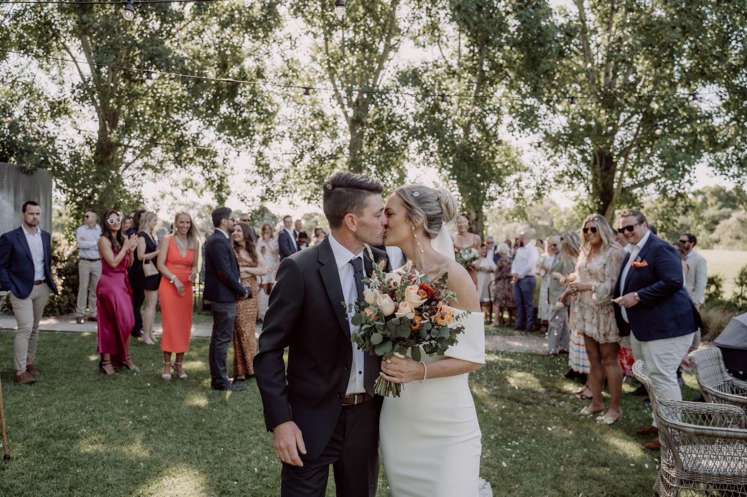 Wedding ceremony outdoors with guests gathered on green lawn under trees while couple shares romantic moment.