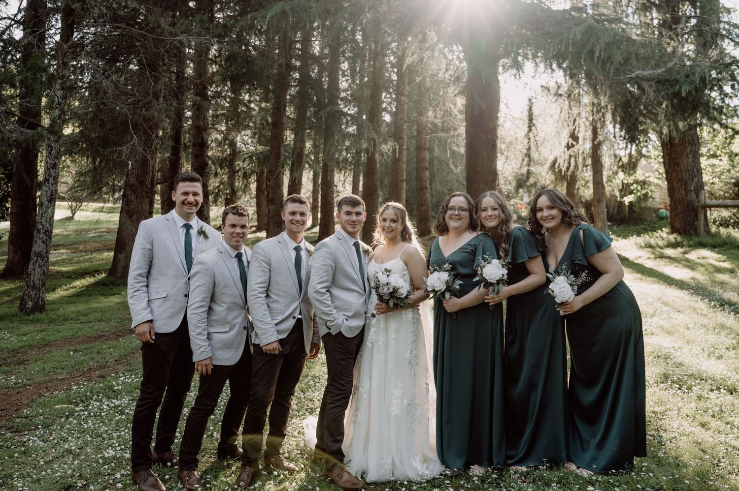 Wedding party poses in a forest setting with sunlight streaming through the trees creating a dreamy atmosphere.