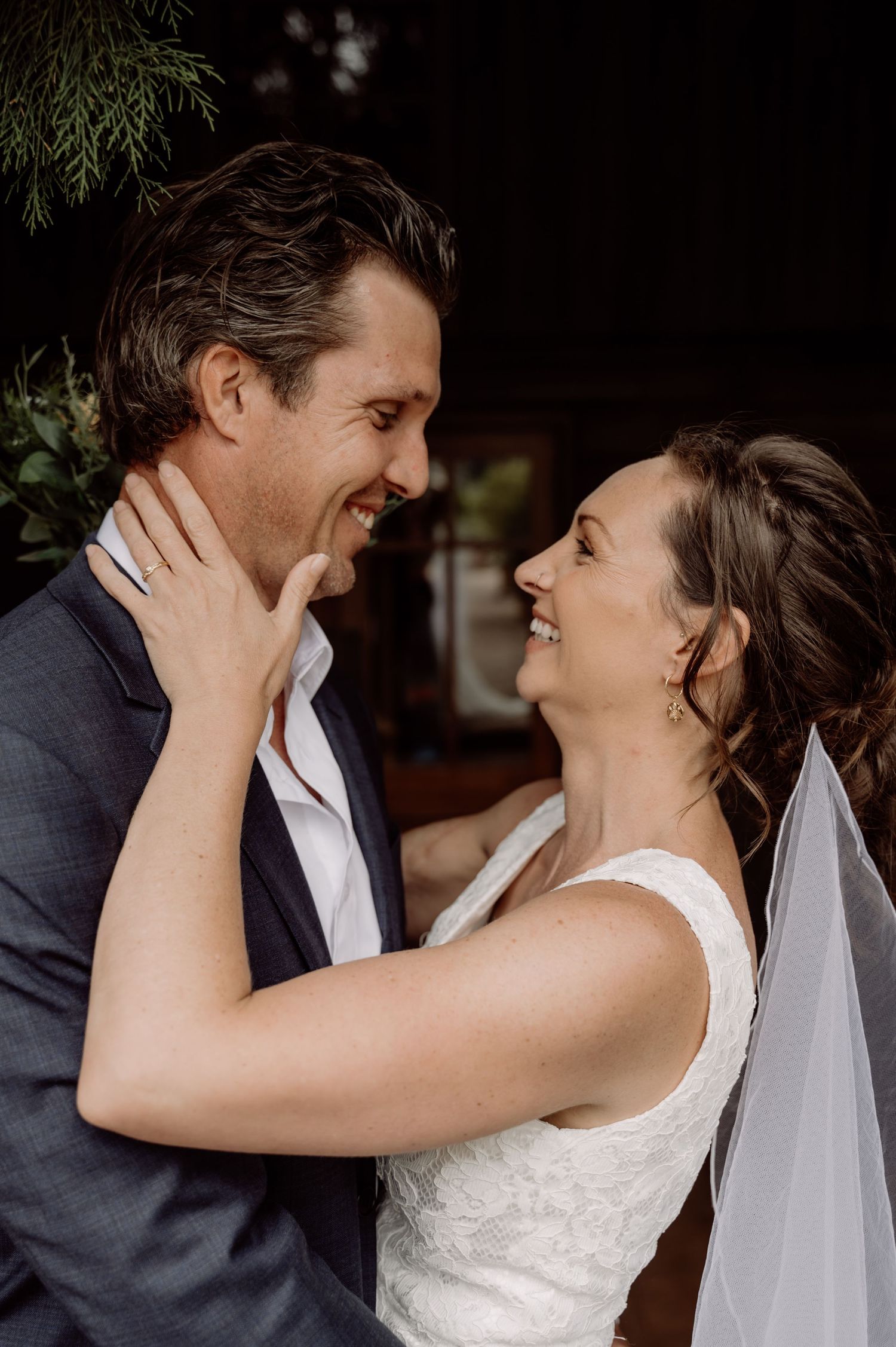 A loving wedding moment shows a couple smiling at each other as the bride in white dress touches her partner's face.