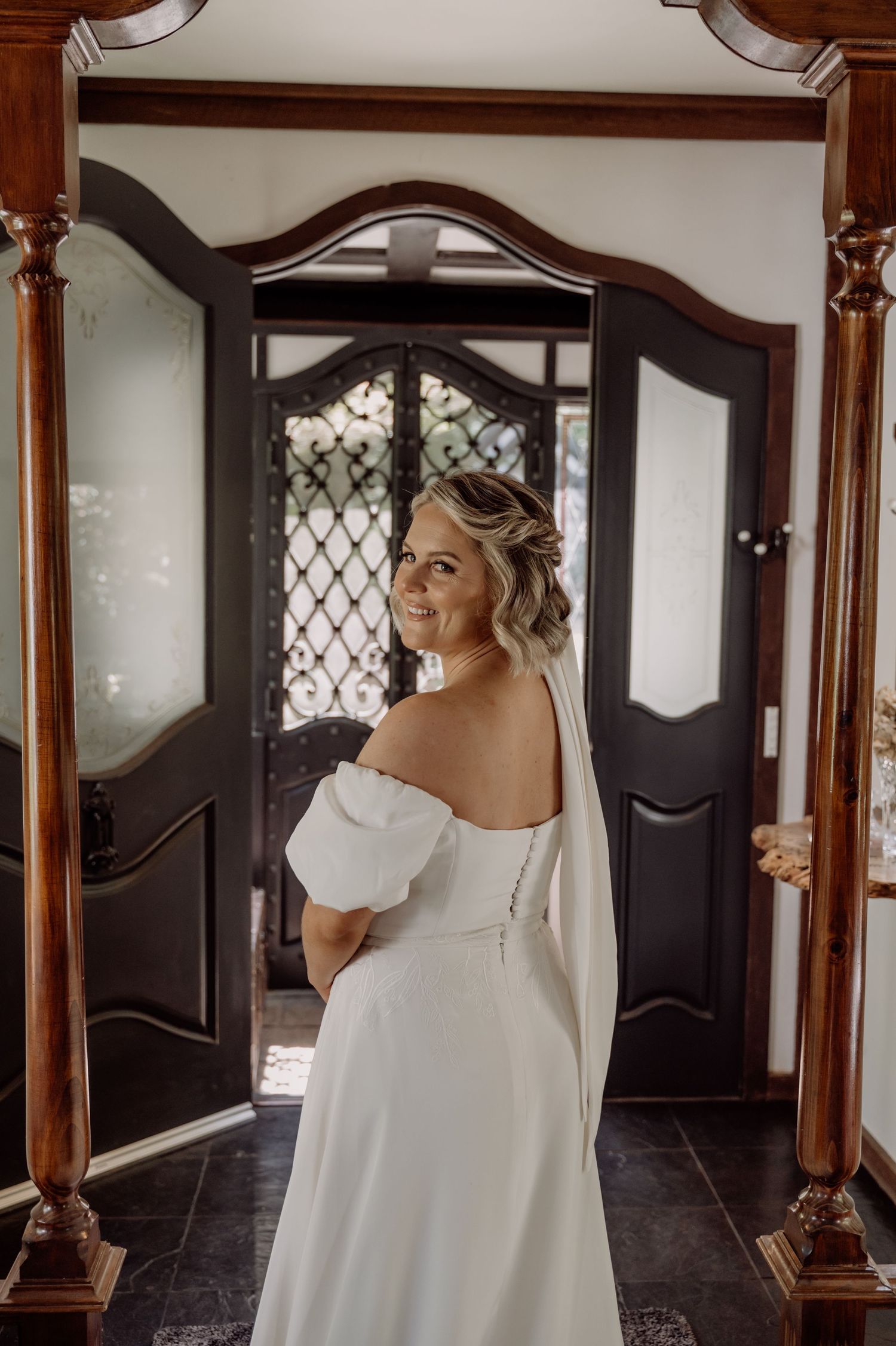 An elegant off-shoulder white wedding gown is shown from behind in a grand entrance hall with ornate wooden doors.