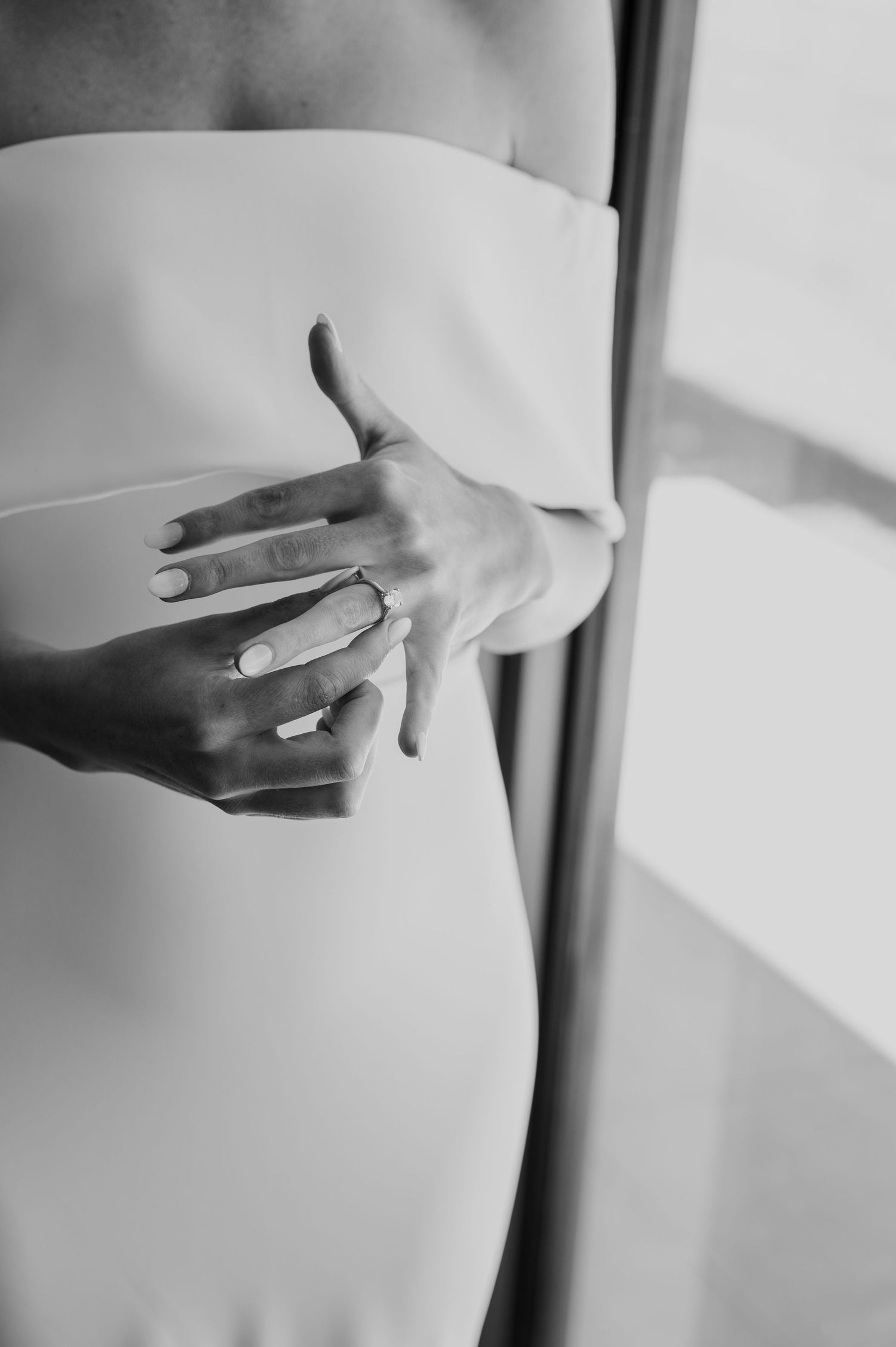 Black and white close-up of a bride's hands gently holding their engagement ring with soft window light.