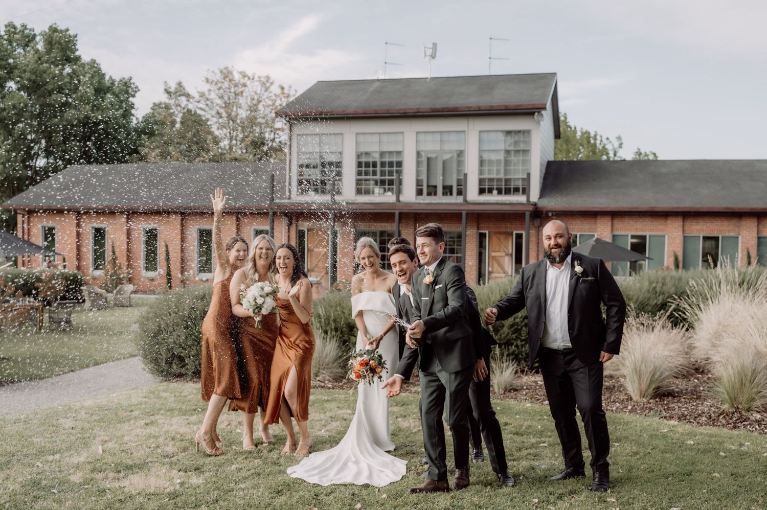 A bridal party group stands together outside a rustic brick and wood venue with ornamental grasses on a sunny day.