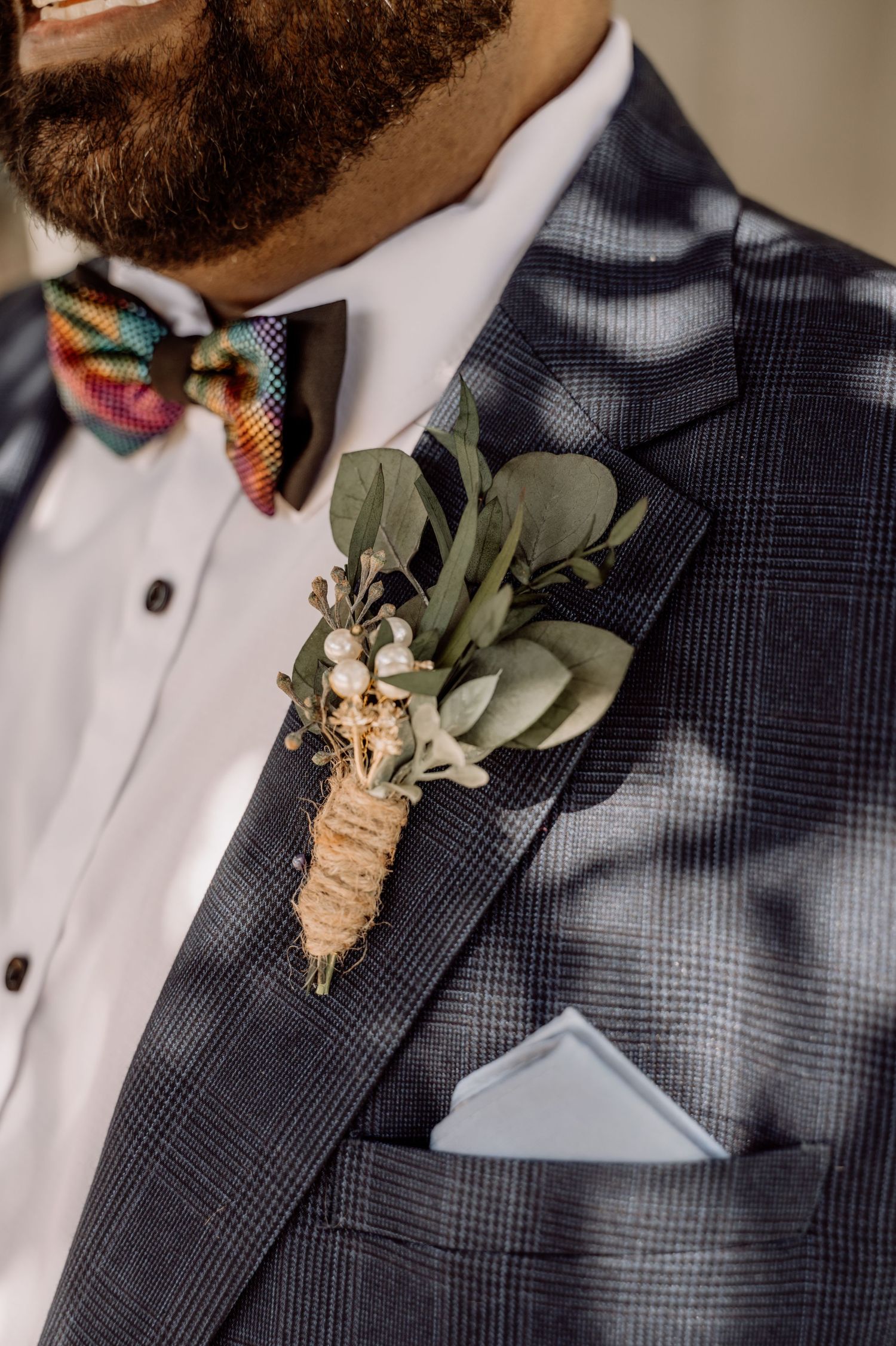 Close-up of groom's attire showing plaid suit jacket, colorful bowtie, and rustic boutonniere with eucalyptus and white flowers.