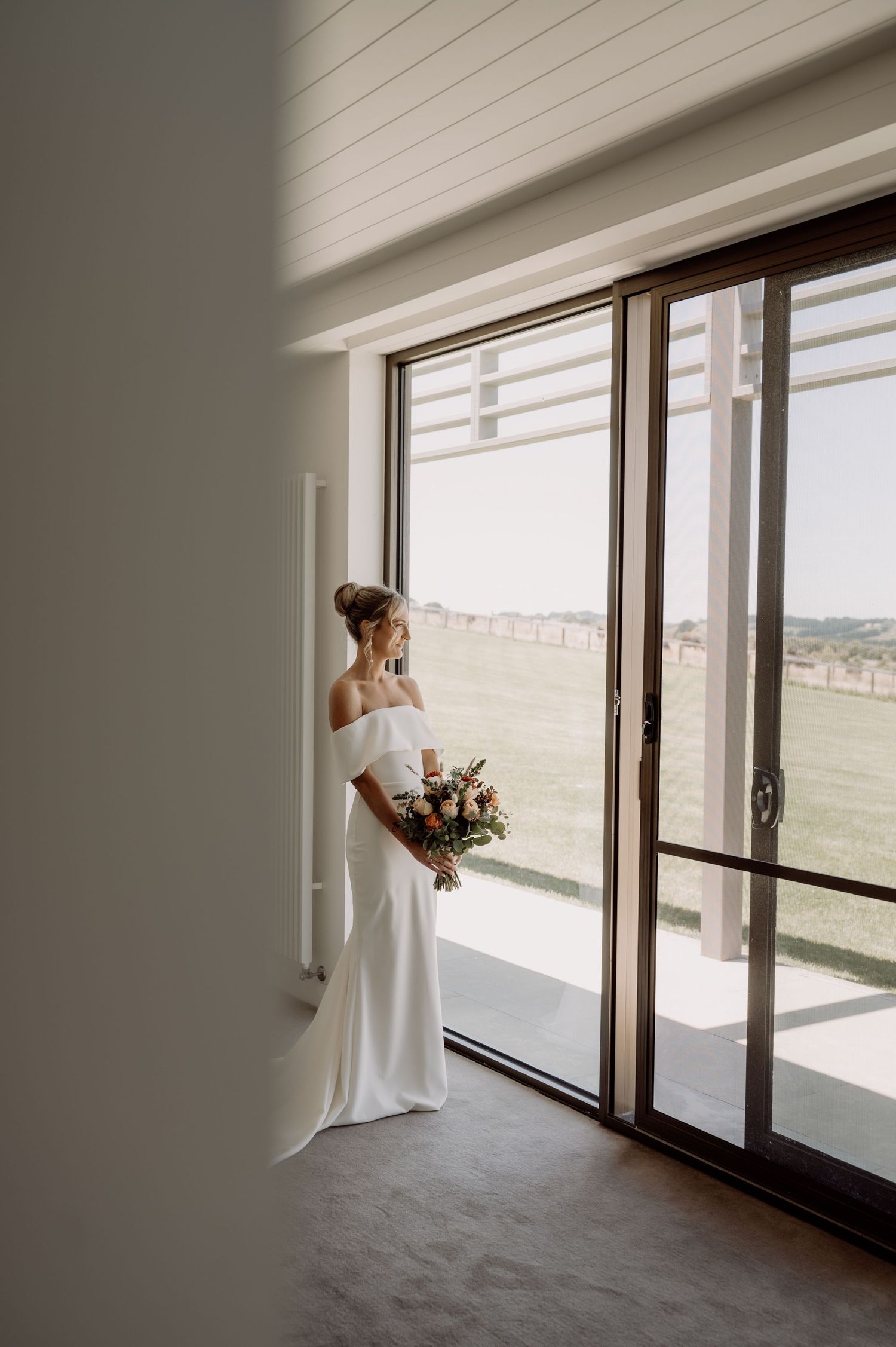 A bridal figure in a white gown stands silhouetted against large floor-to-ceiling windows in a minimalist interior space.
