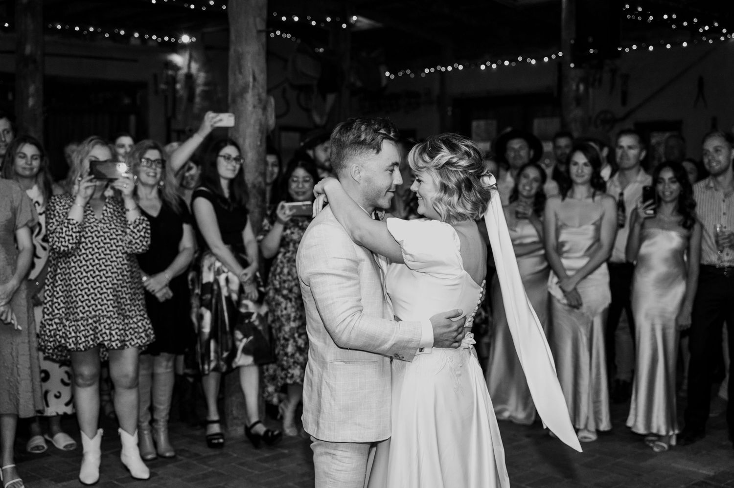 A couple shares their first dance at a wedding reception surrounded by guests in a rustic venue with string lights.
