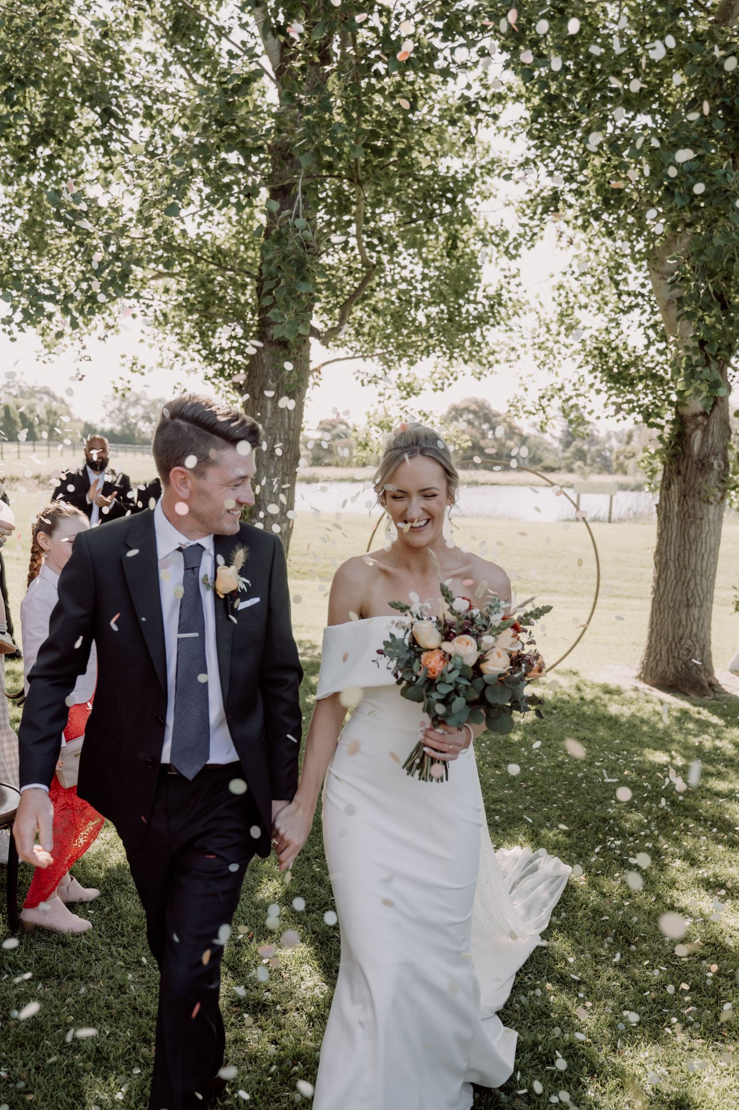 A newly wed couple walking together outdoors among trees on a bright sunny day, with confetti falling through the air.