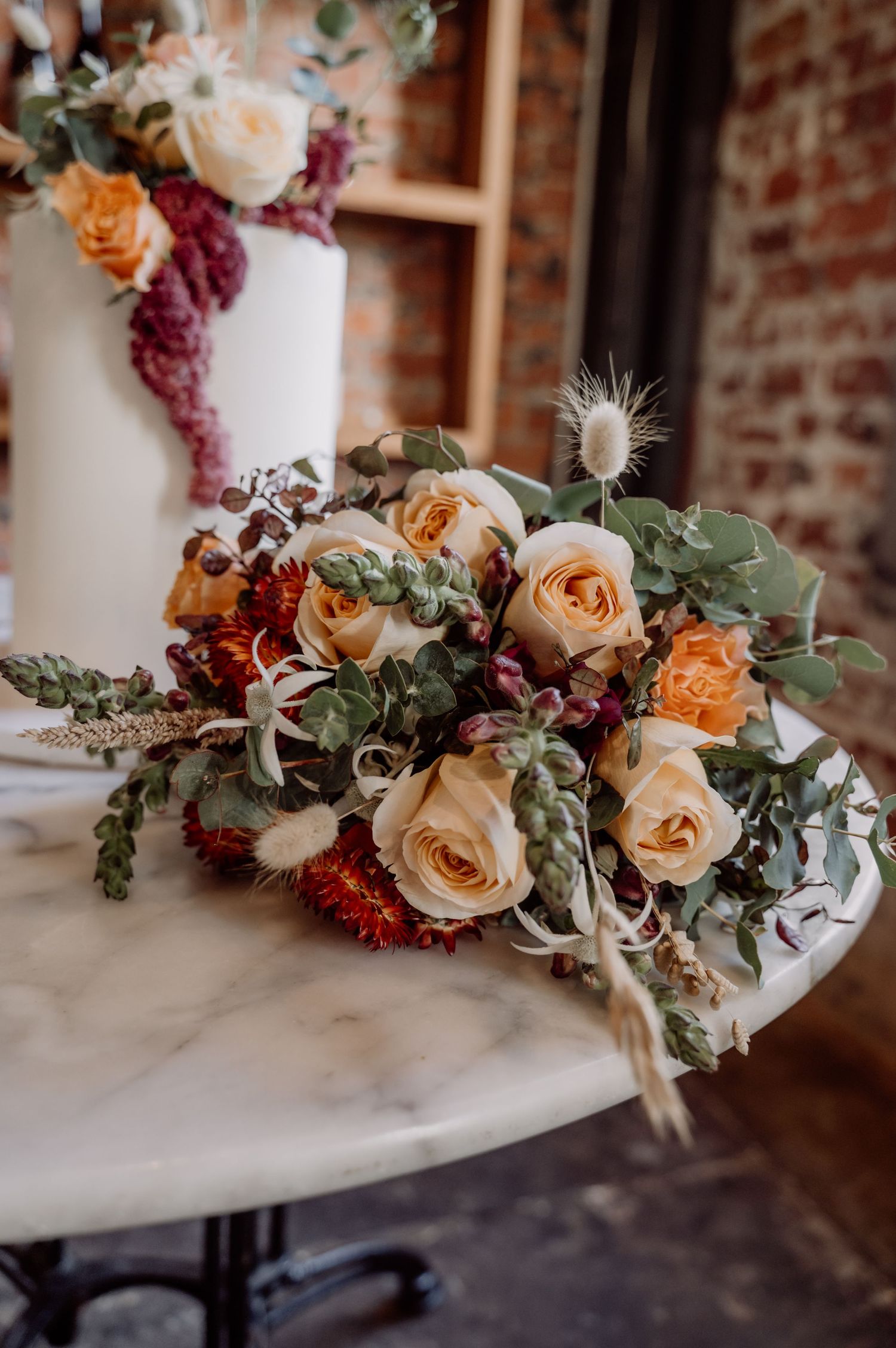 Beautiful peach roses and burgundy florals arranged with eucalyptus leaves on a marble table against exposed brick wall.
