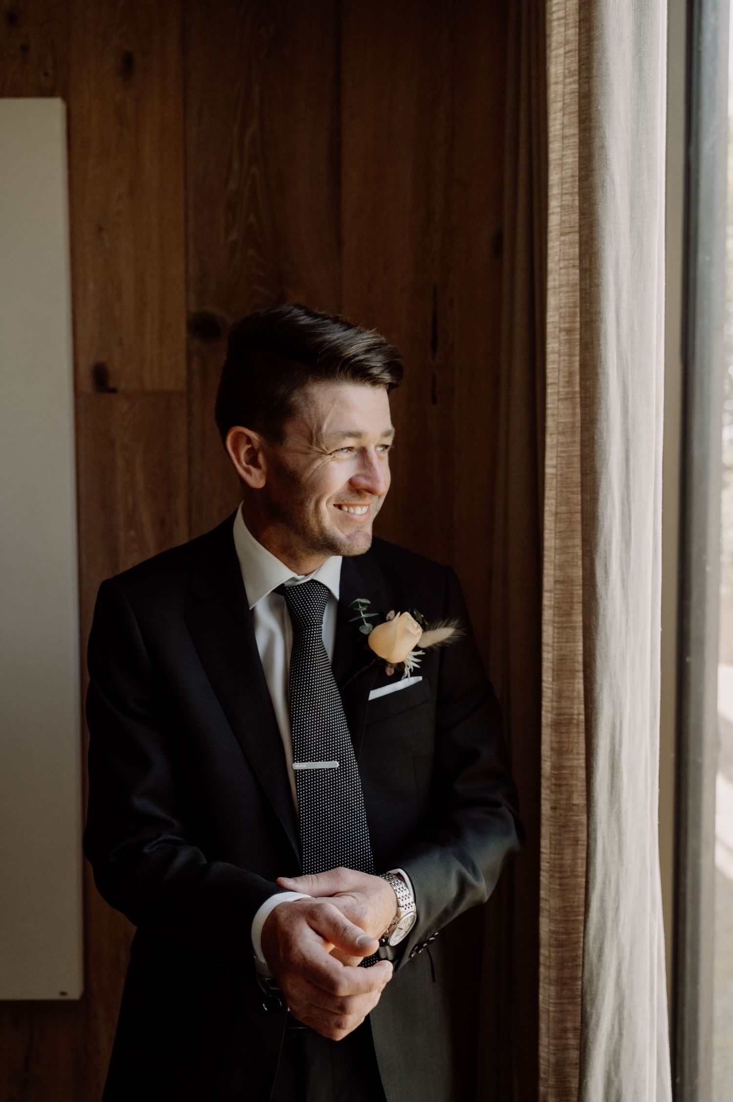 A groom in a black suit with a boutonniere smiles while looking out a window in warm natural light.