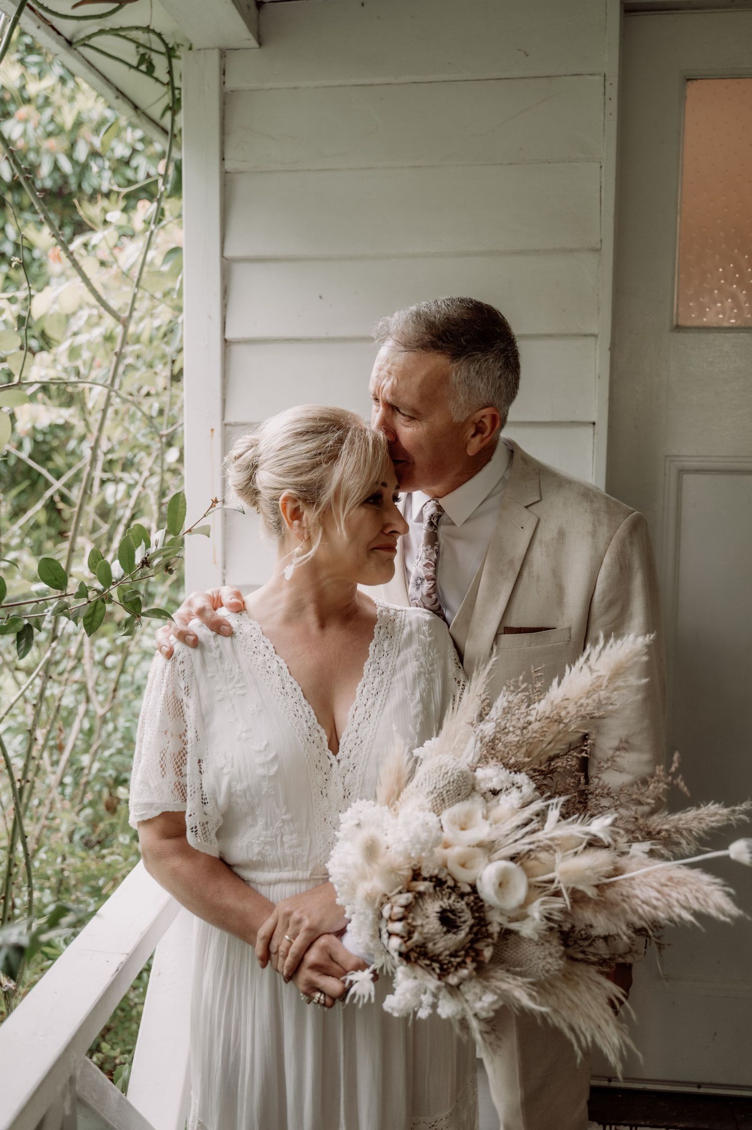 A couple in wedding attire share an intimate moment on a porch, with the bride holding a romantic dried floral bouquet.
