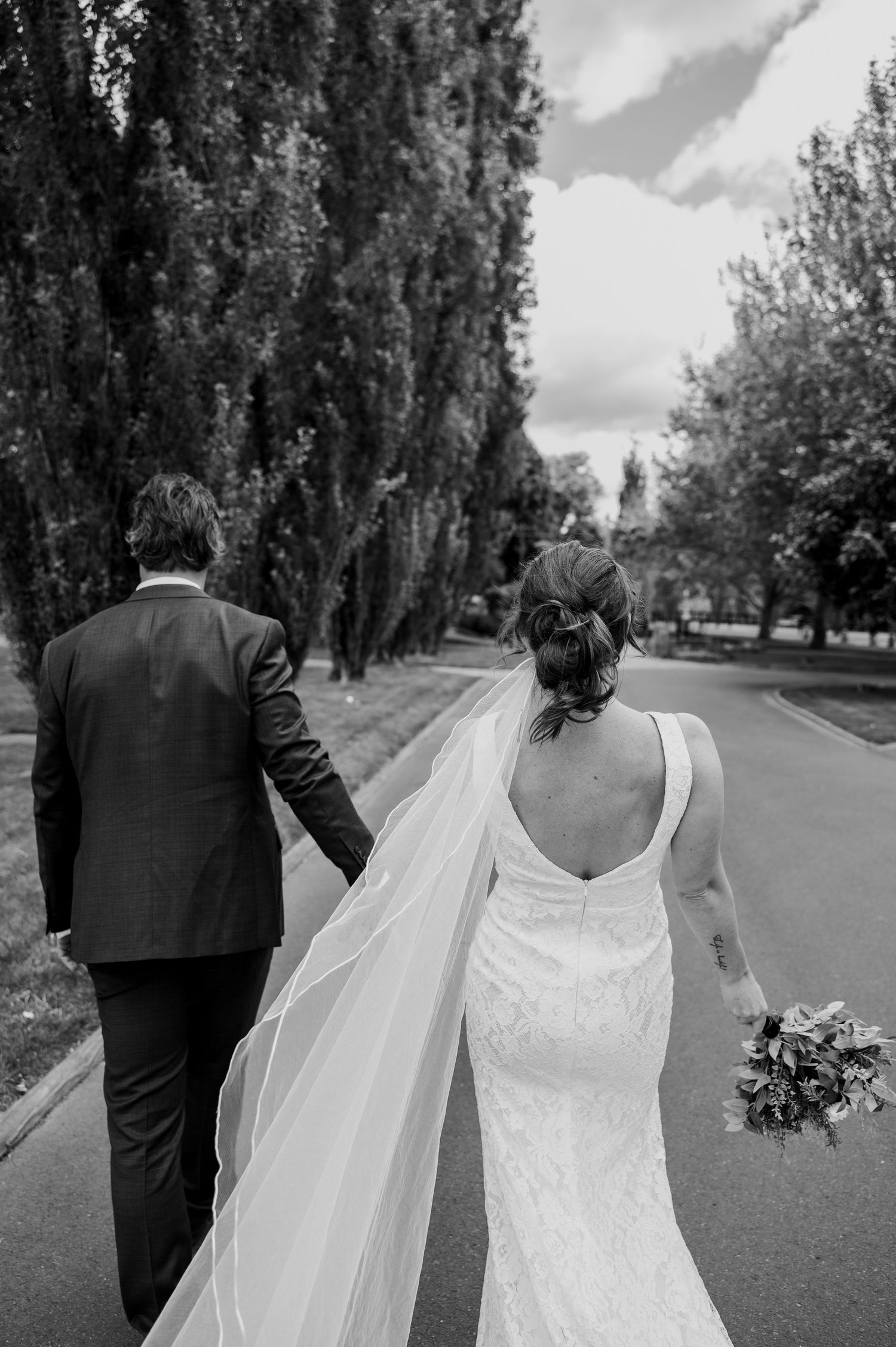 Black and white wedding photo of couple walking down tree-lined road with bride in white gown holding bouquet.