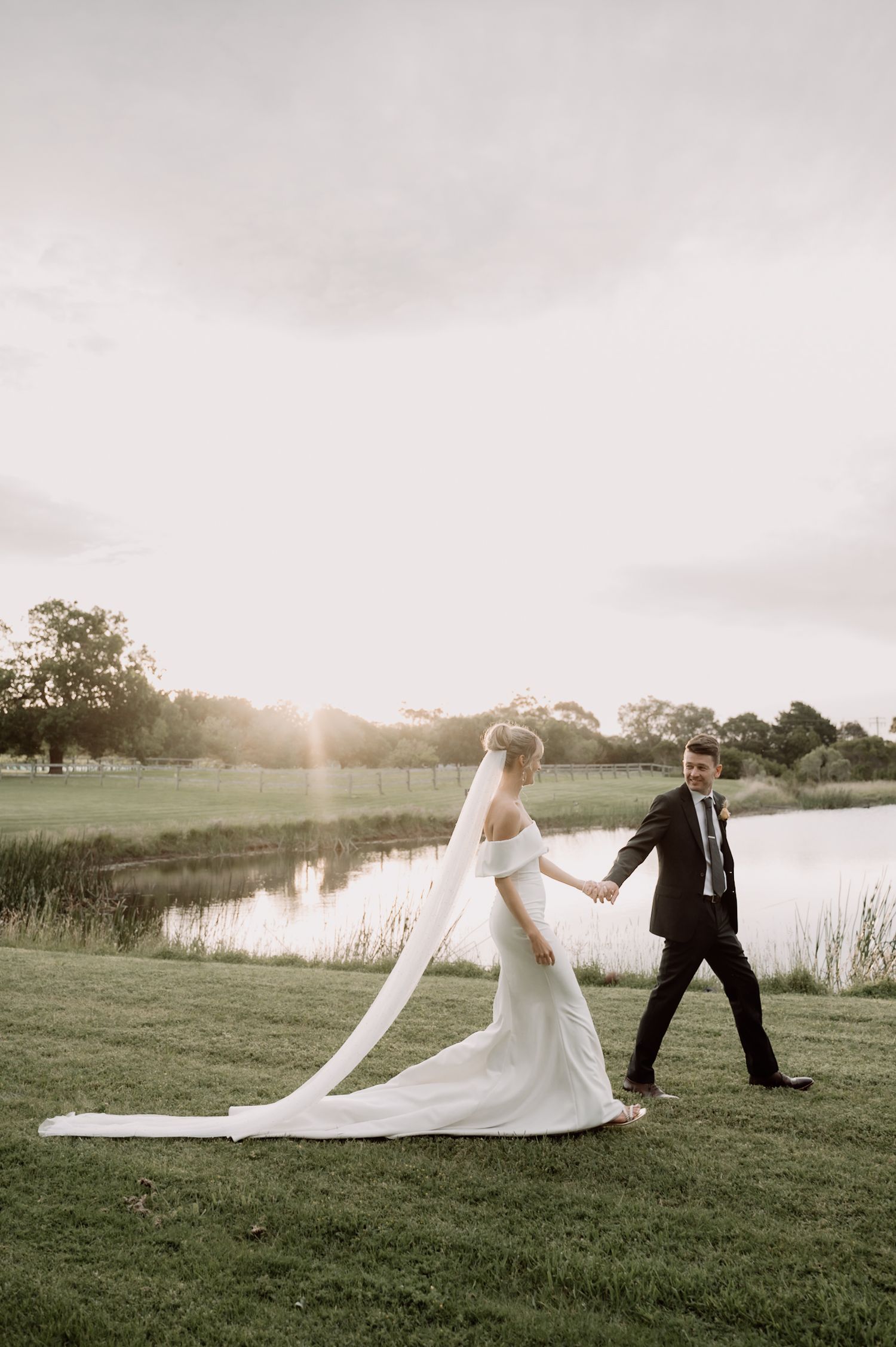 A couple in wedding attire walks along a scenic lakeside at sunset with a flowing white veil trailing behind them.