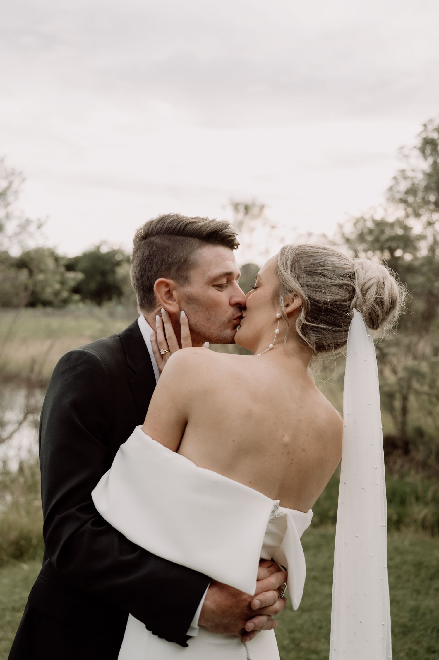 A romantic wedding photo of a couple sharing a kiss outdoors in a natural setting with the bride wearing a white dress and veil.