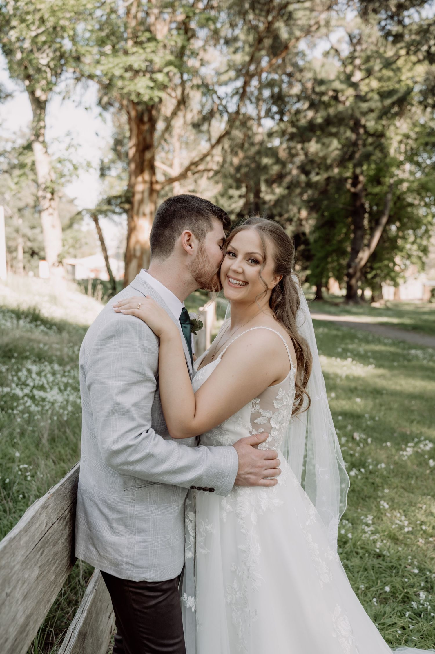 A romantic wedding couple embraces outdoors in a scenic garden setting with sunlit trees and wildflowers in the background.