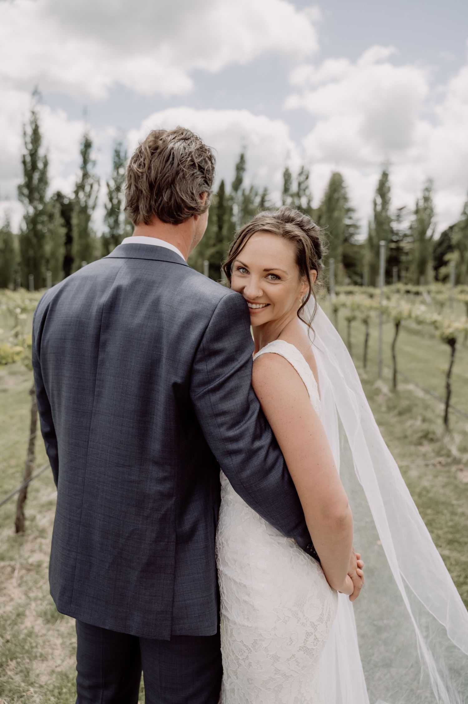 Newlywed couple poses outdoors with tall trees in background while bride looks back at camera wearing flowing veil.