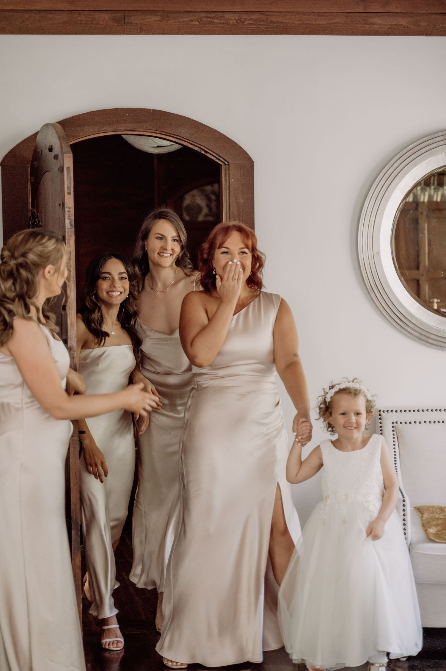 Emotional wedding party gathering in shades of white and cream dresses in a room with circular mirrors on the wall.