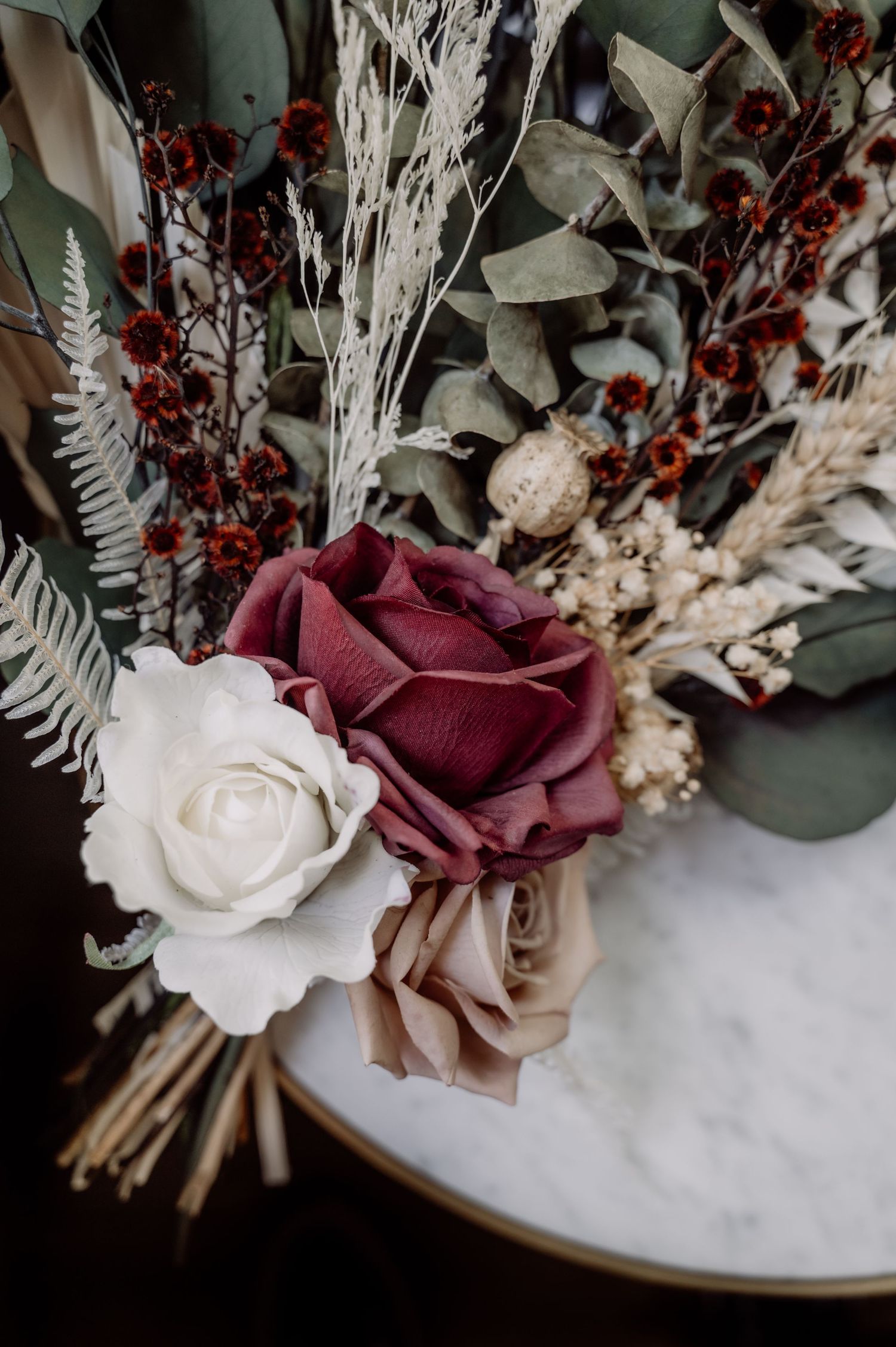 A romantic dried floral arrangement with burgundy and white roses, eucalyptus leaves, and white dried grasses on marble.