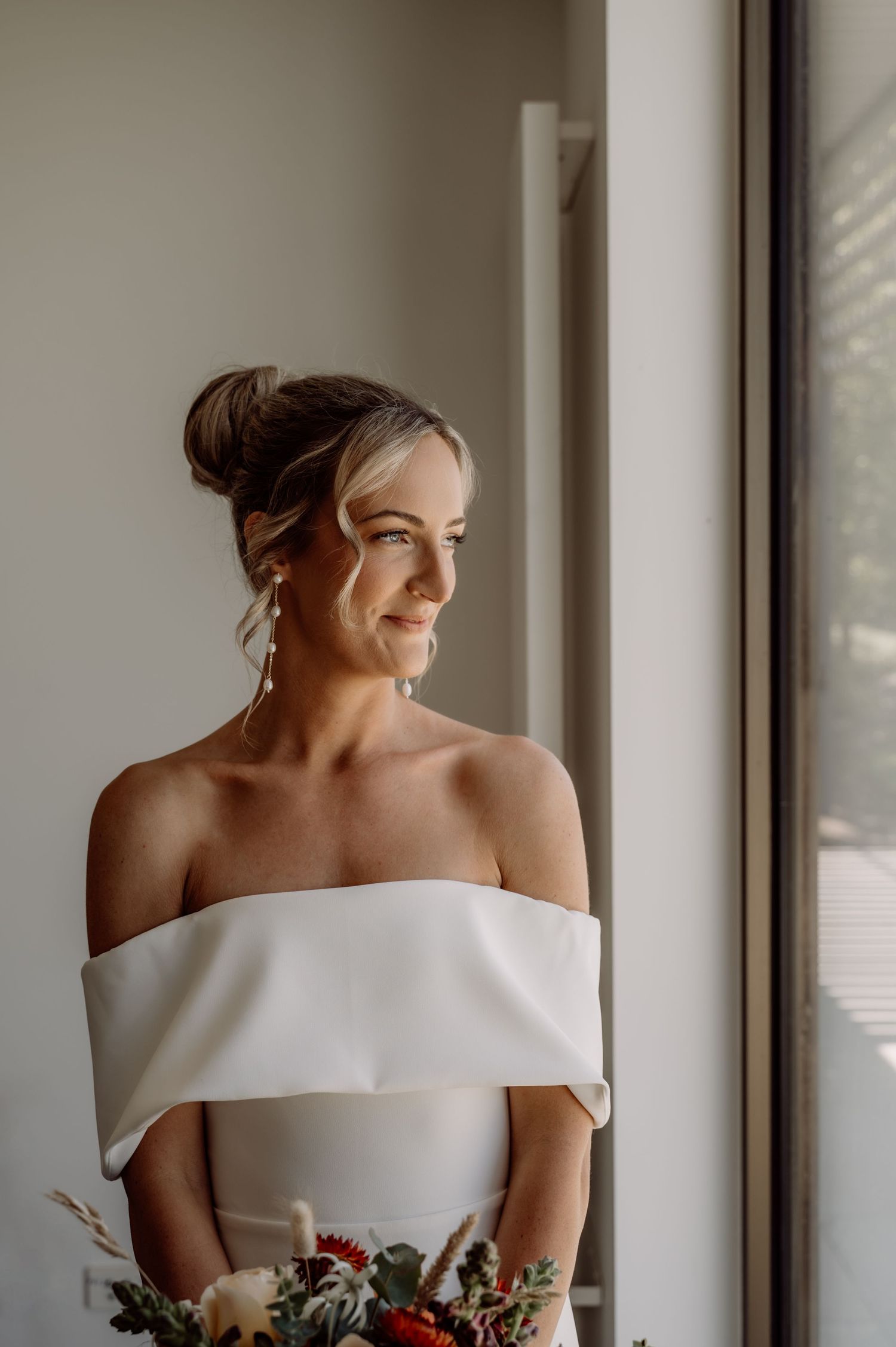 Bride in elegant off-shoulder white wedding dress poses by window in a series of romantic minimalist portraits.