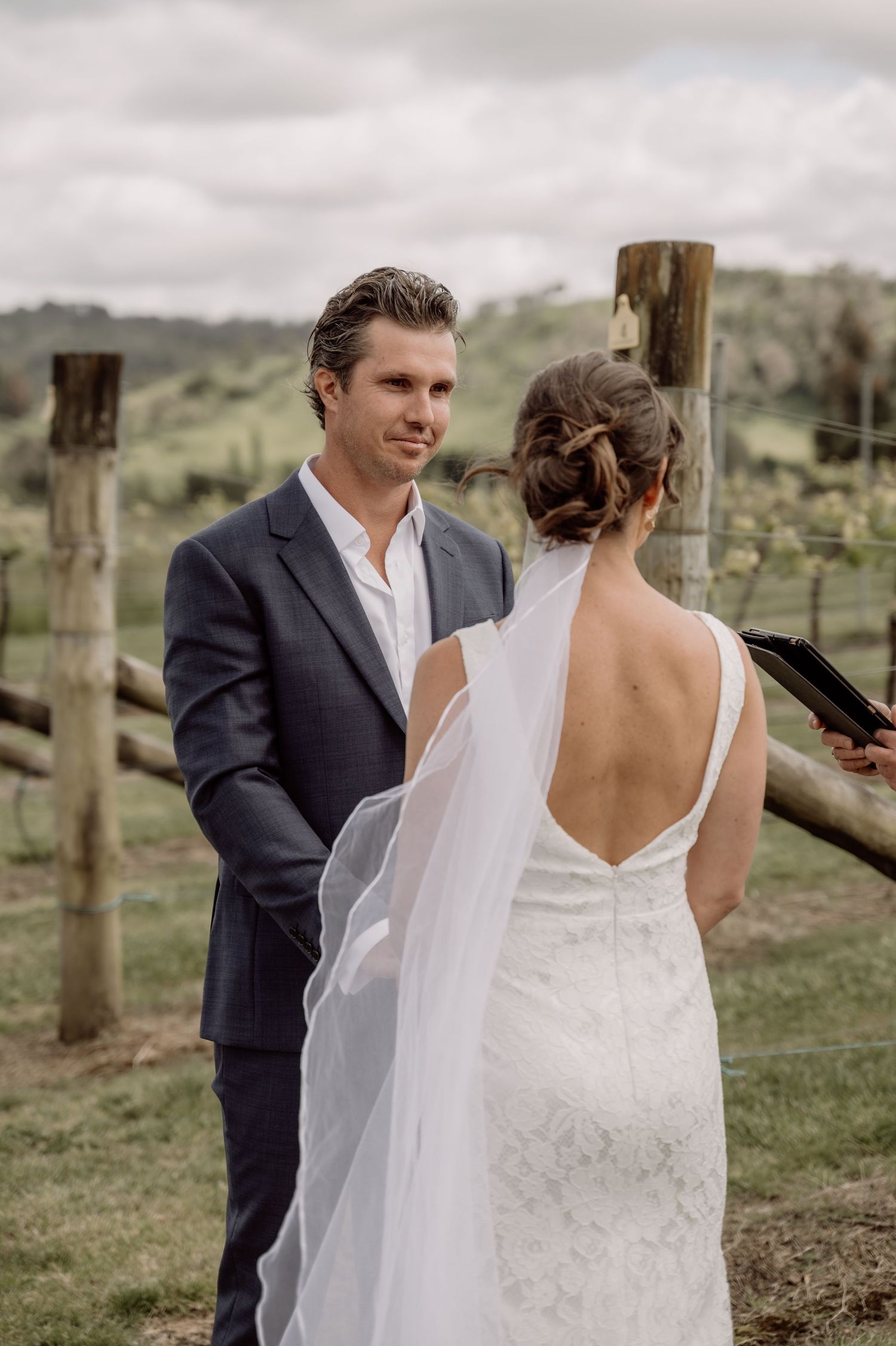 A couple in wedding attire share an intimate candid moment in a rustic vineyard setting with wooden posts.