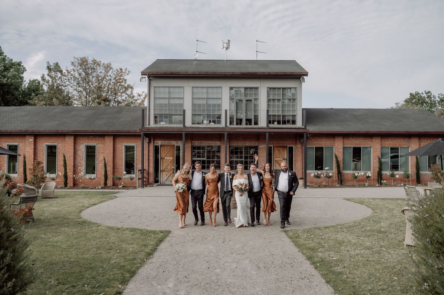 Wedding party poses together on a curved path outside a modern brick building with glass atrium on a cloudy day.