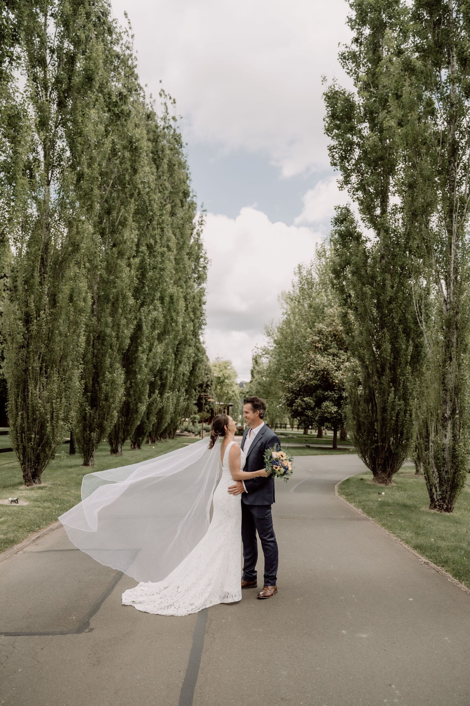 A bride and groom walk down a tree-lined path in a romantic outdoor setting during their wedding day photoshoot.