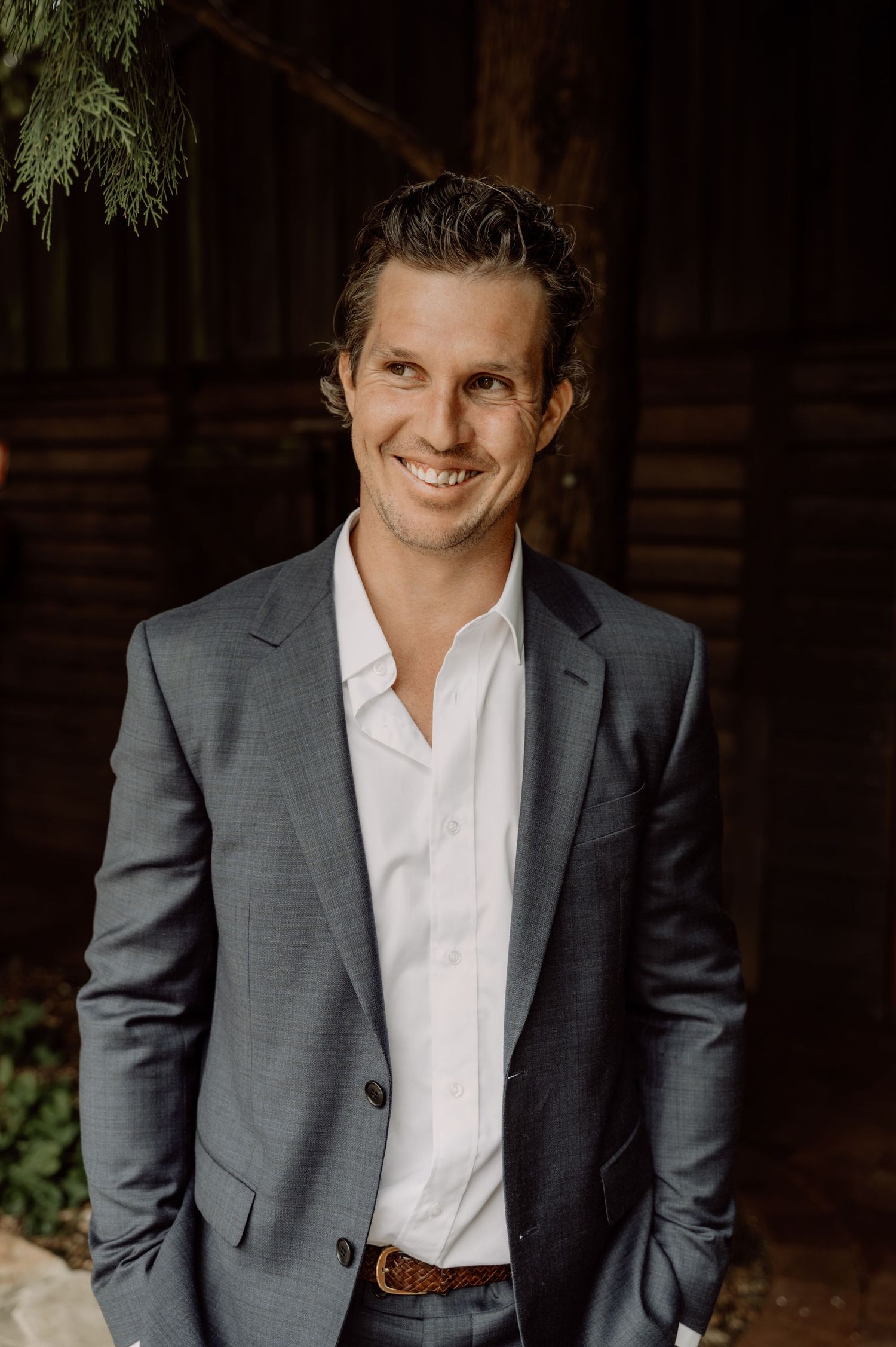 Professional wedding portrait of a groom in a gray suit jacket and white shirt standing against a rustic wooden wall background.