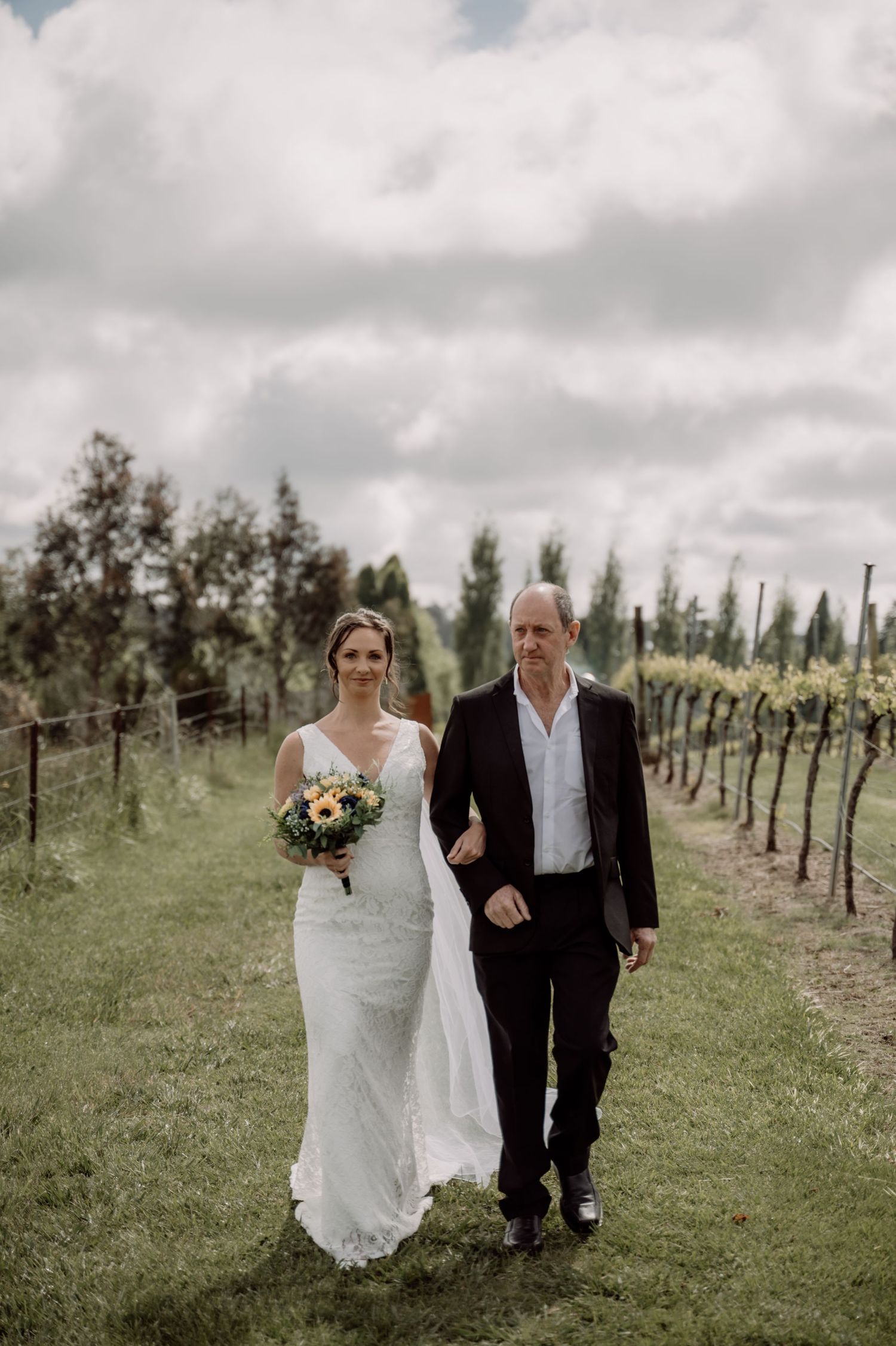 A wedding couple walking arm in arm down a grassy path between rows of grapevines at a vineyard under cloudy skies.