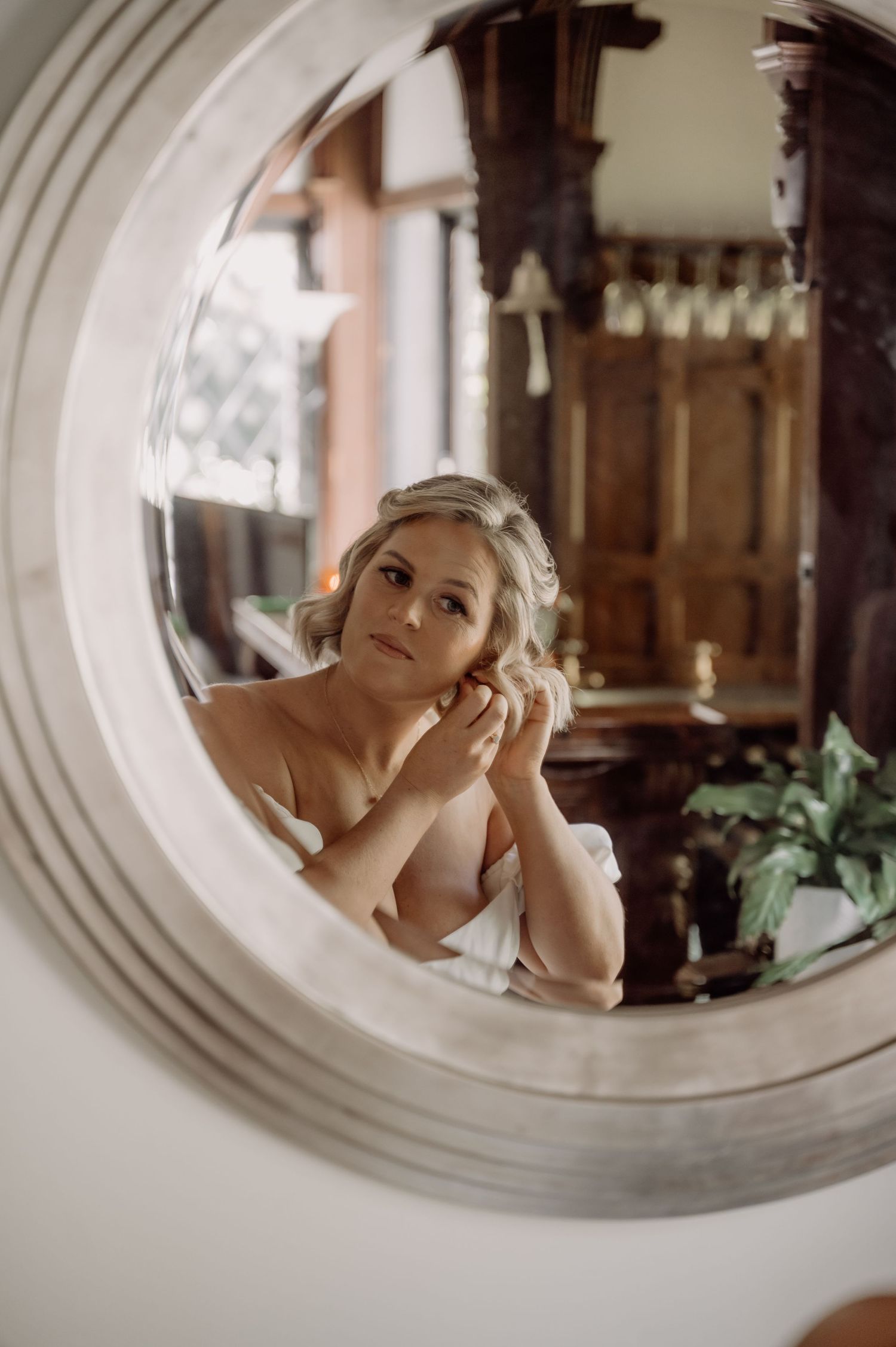 A reflection in a round mirror shows a romantic bridal preparation scene with soft lighting and antique wooden decor.