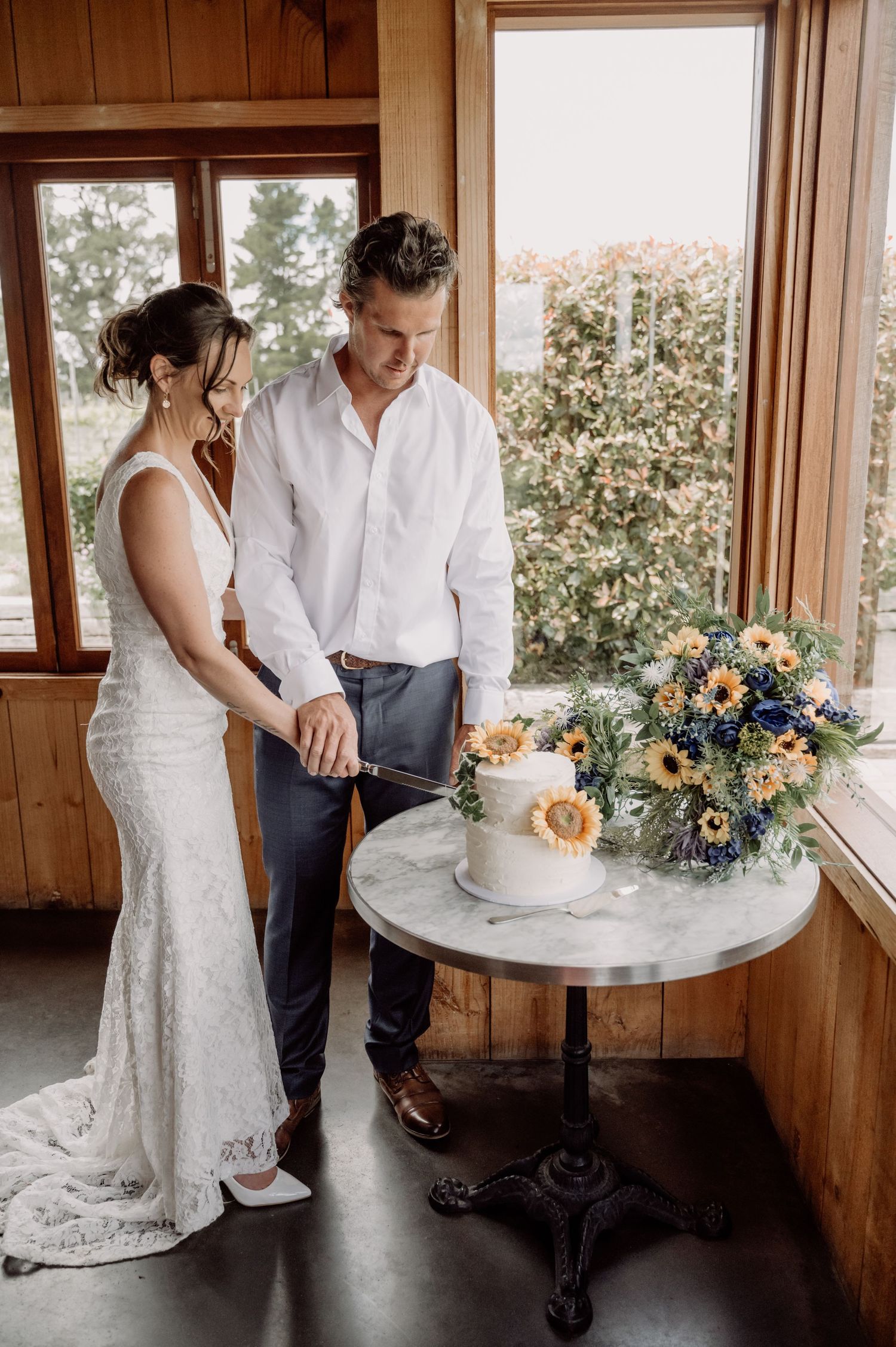 A couple cuts their wedding cake together on a marble-topped table with a beautiful floral arrangement by the window.