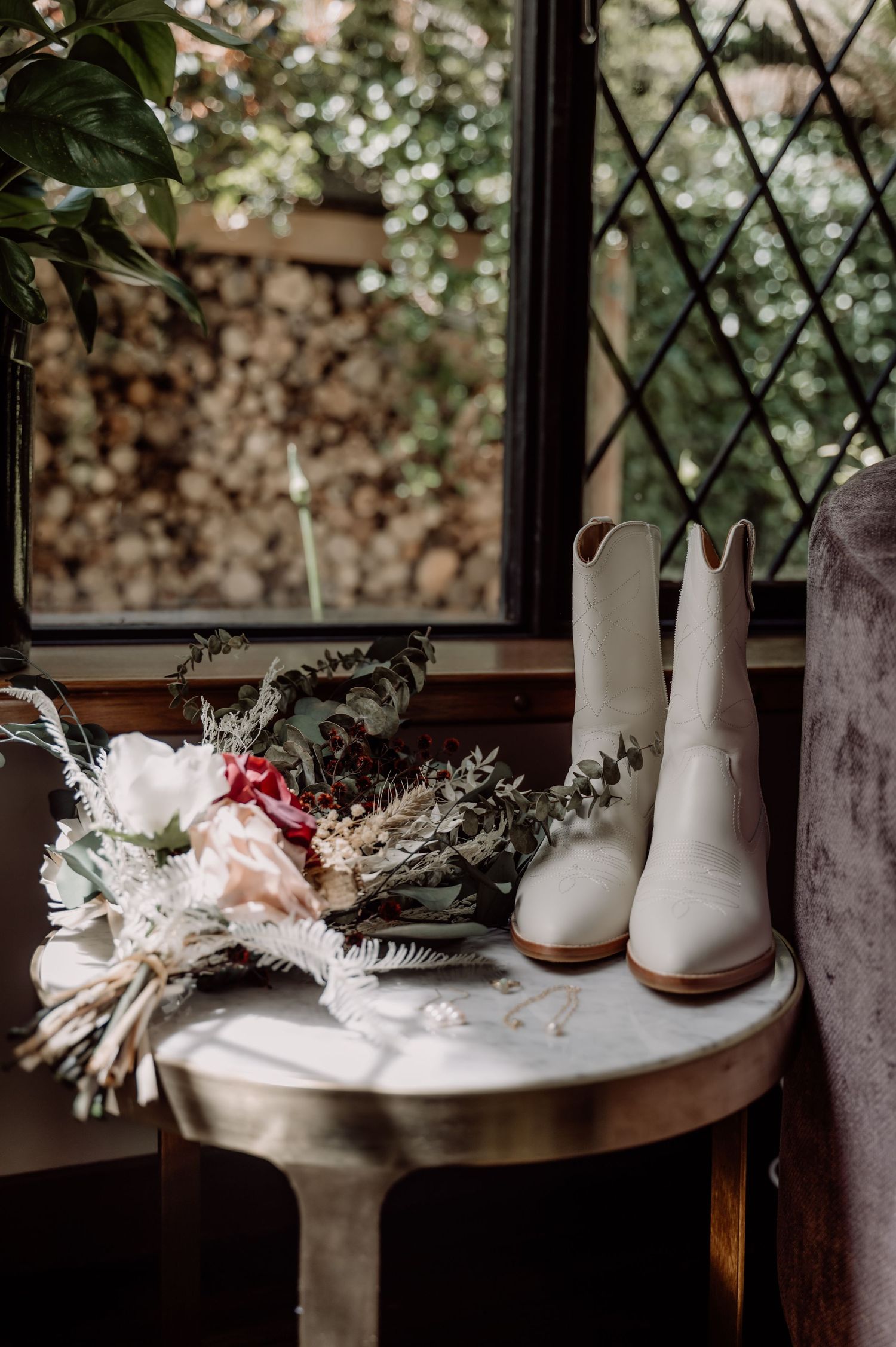 White cowboy boots and a delicate bridal bouquet with dried flowers sit on a rustic table by a window.