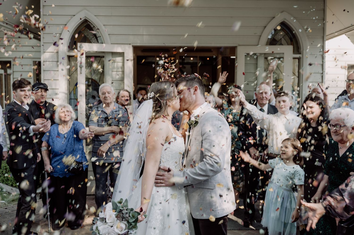 Joyful wedding celebration outside historic house entrance with confetti floating through the air during spring ceremony.