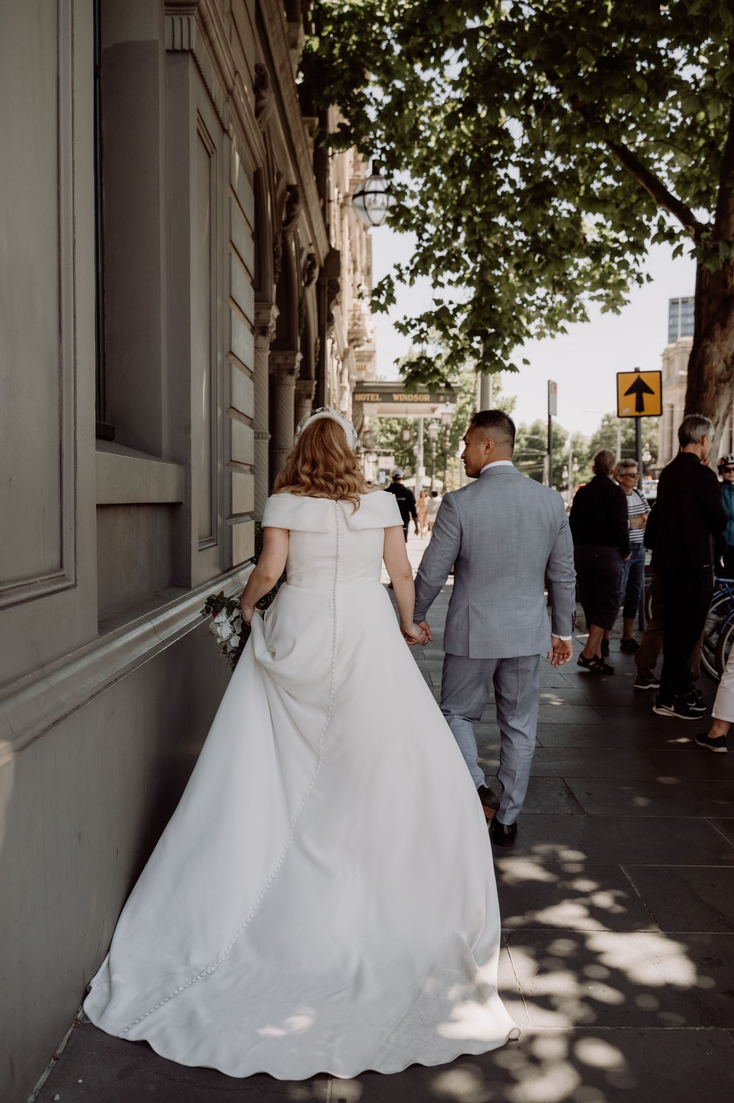 A couple in wedding attire walks along a city sidewalk under dappled tree shadows.
