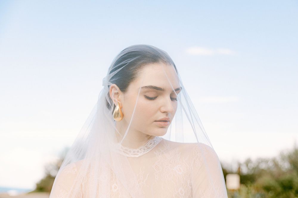 Ethereal bridal portrait with delicate veil against soft blue sky backdrop.