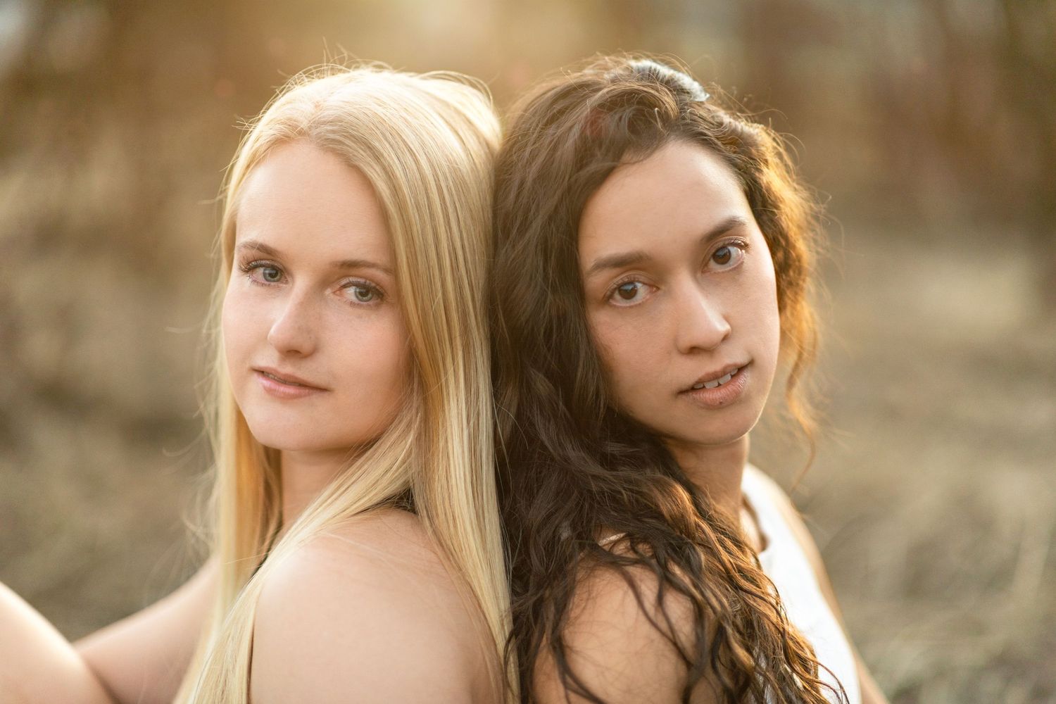 two girls back to back sitting leaning their heads together and looking at camera.