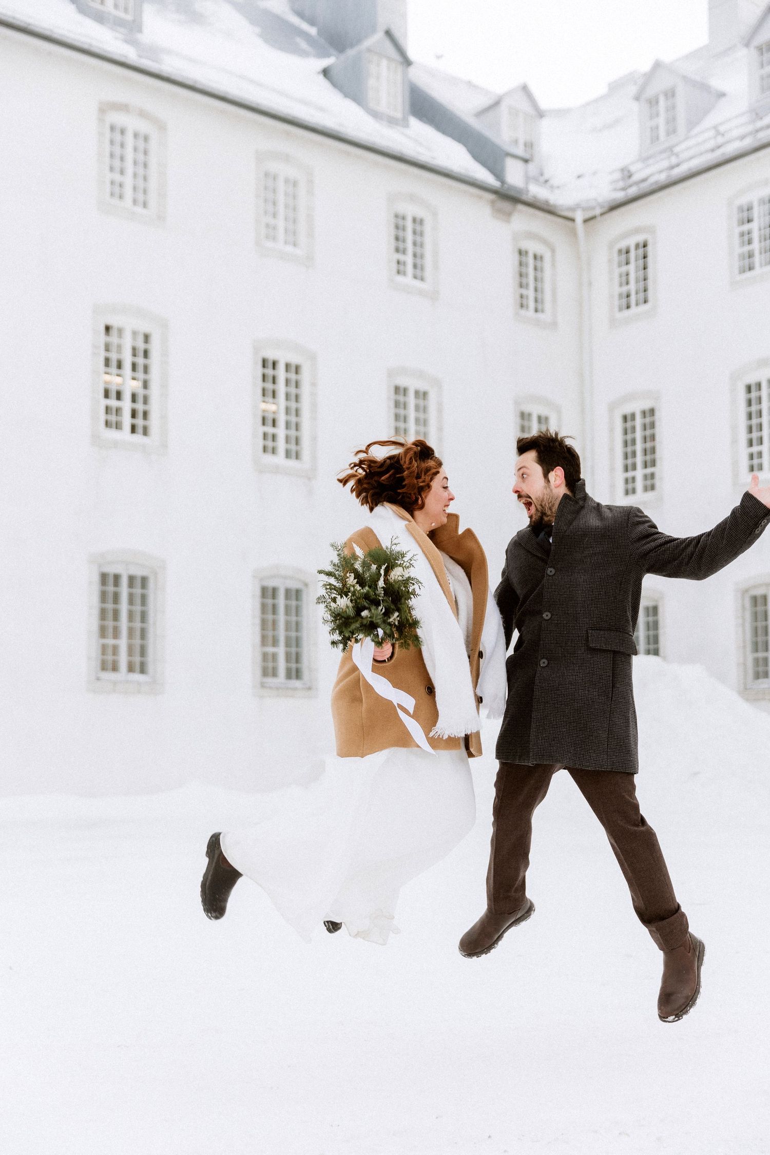 A joyful couple jumps in the snow during their wedding photoshoot in front of a white historic building.