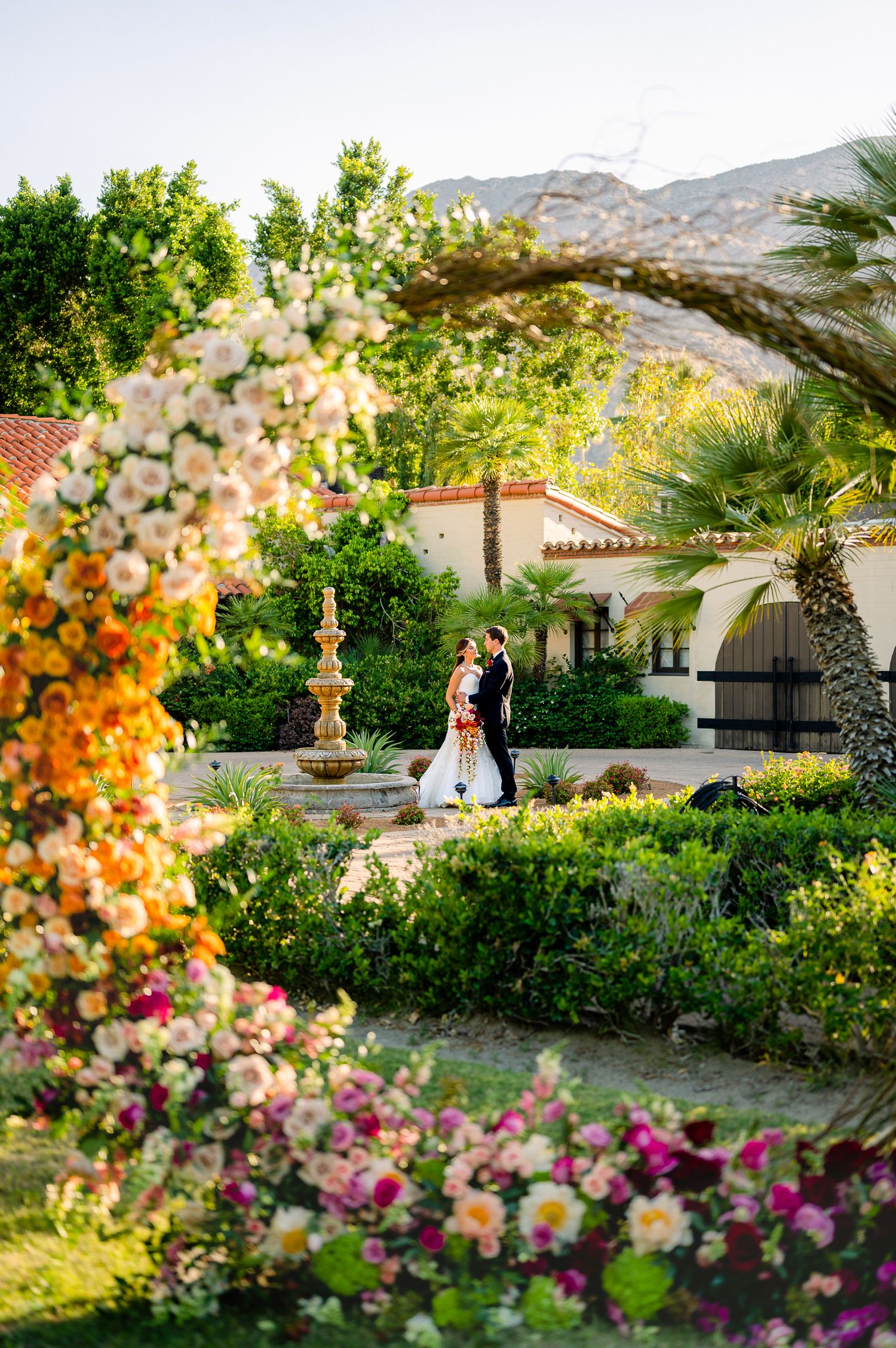A garden wedding scene framed by a floral arch with Spanish architecture in the background.