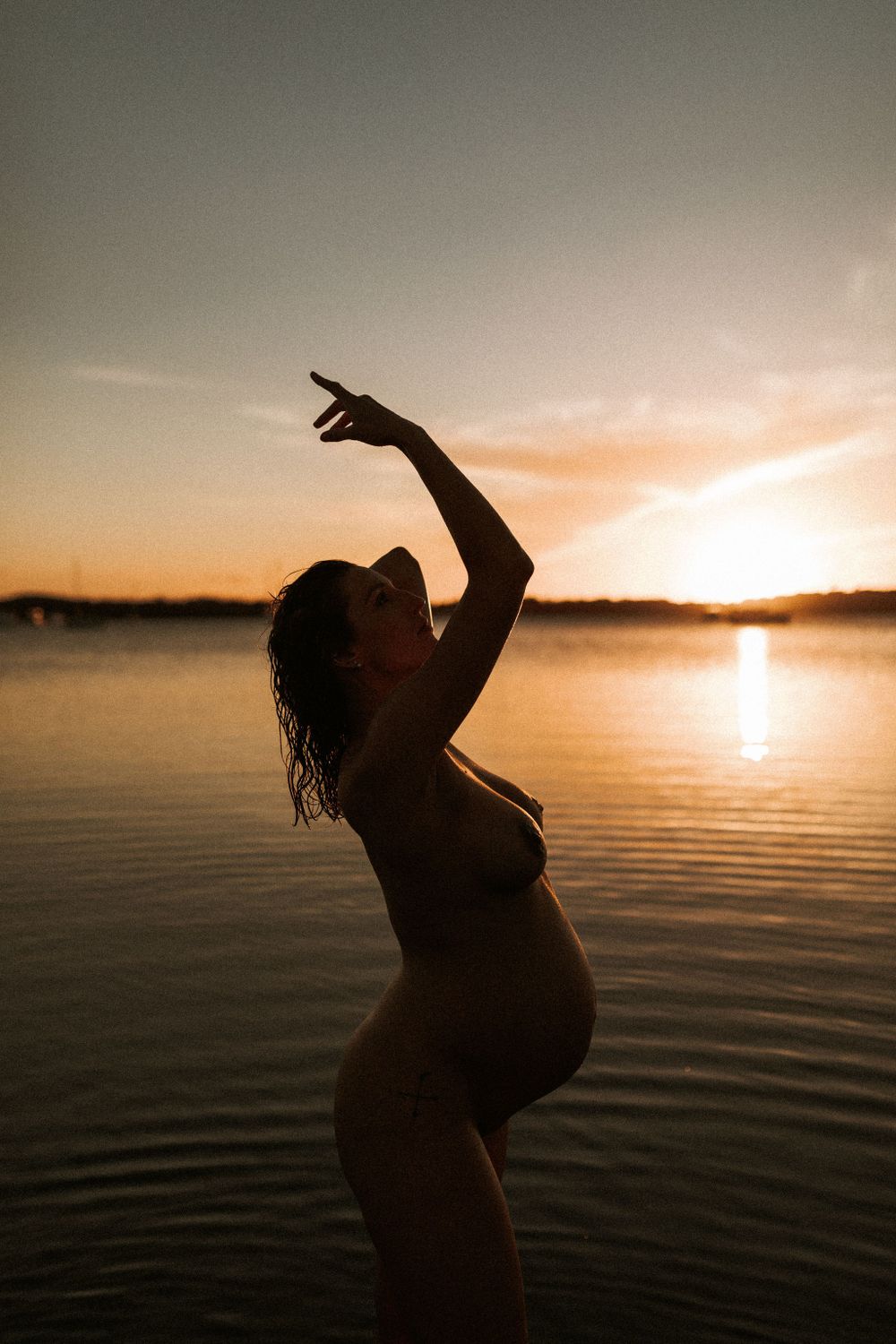 Silhouette of pregnant woman dancing by water at sunset.