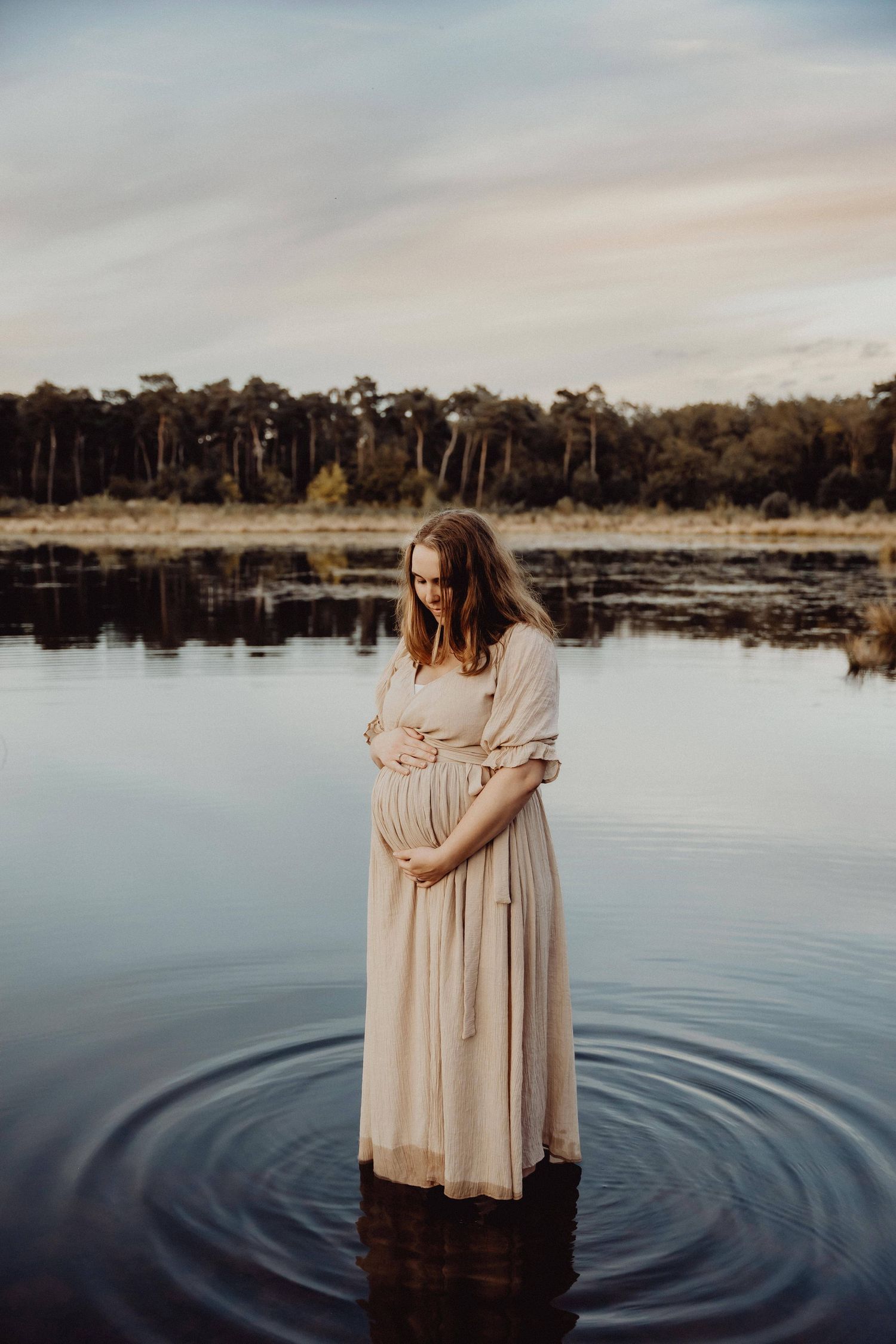 Person in light dress standing in calm lake water creating ripples at sunset.