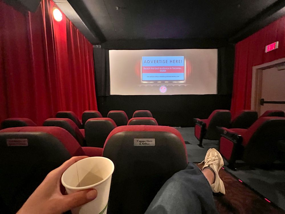 Movie theater interior showing empty red seats and screen with someone holding a drink.