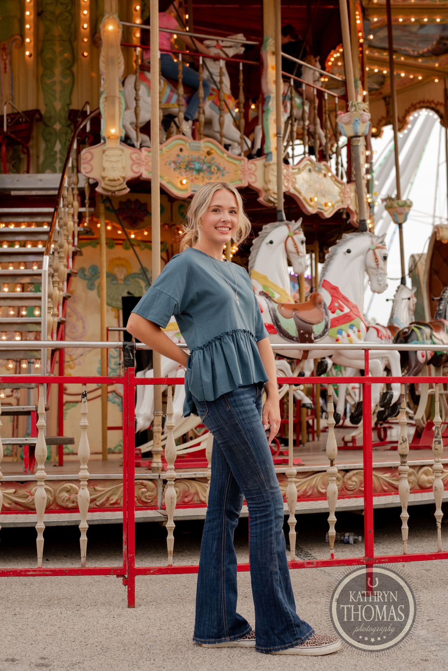 Person in blue ruffled top and jeans standing in front of nostalgic carousel horses.