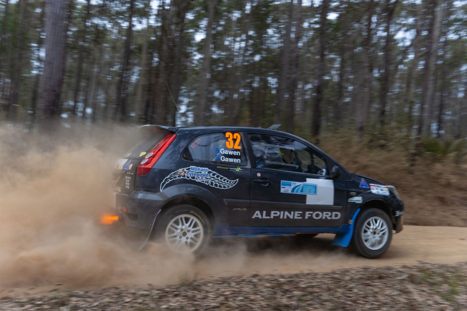 Rally car kicks up dust while racing through forest track.