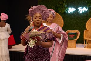 Guest in burgundy dress and headwrap checking phone at Nigerian wedding reception.
