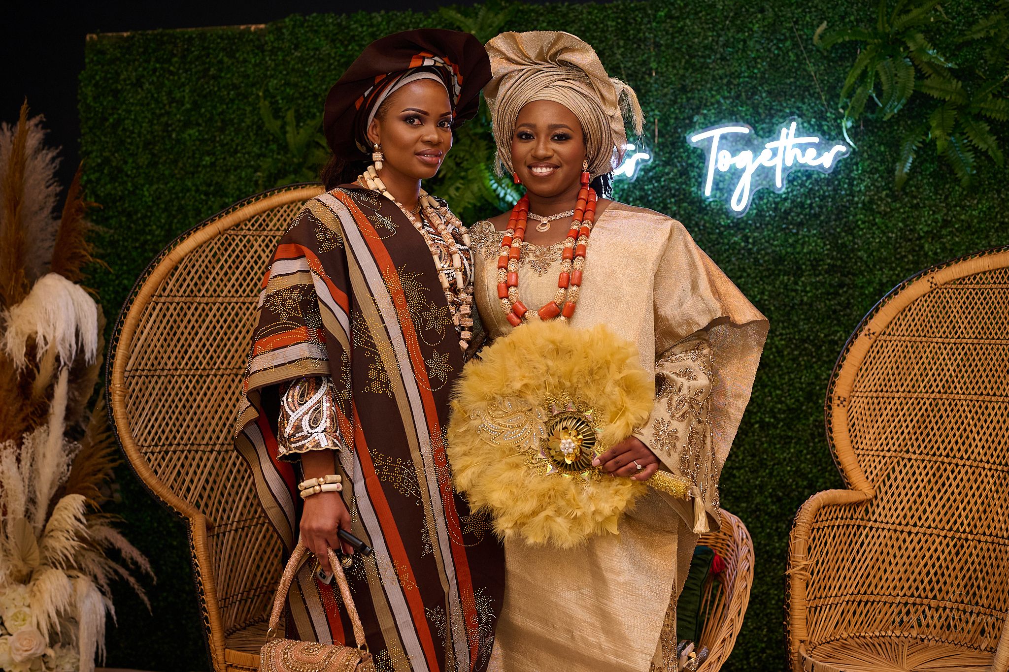 Two guests in traditional Nigerian formal wear posing by wicker chairs and neon sign.
