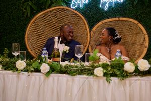 Wedding couple seated at decorated reception table with peacock chairs and floral arrangements.