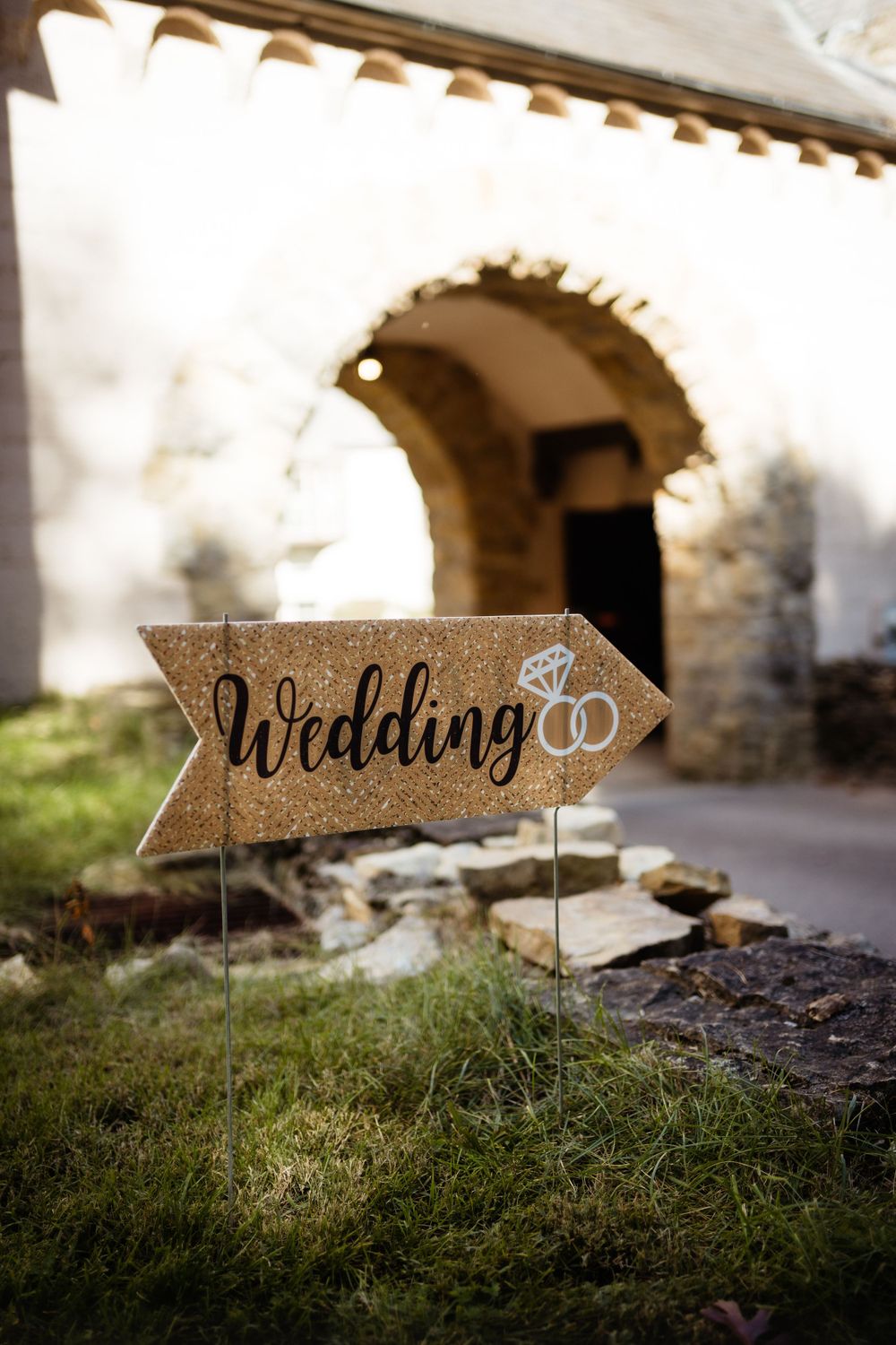 A rustic wooden wedding directional sign with interlocking rings decoration stands in front of a stone archway.