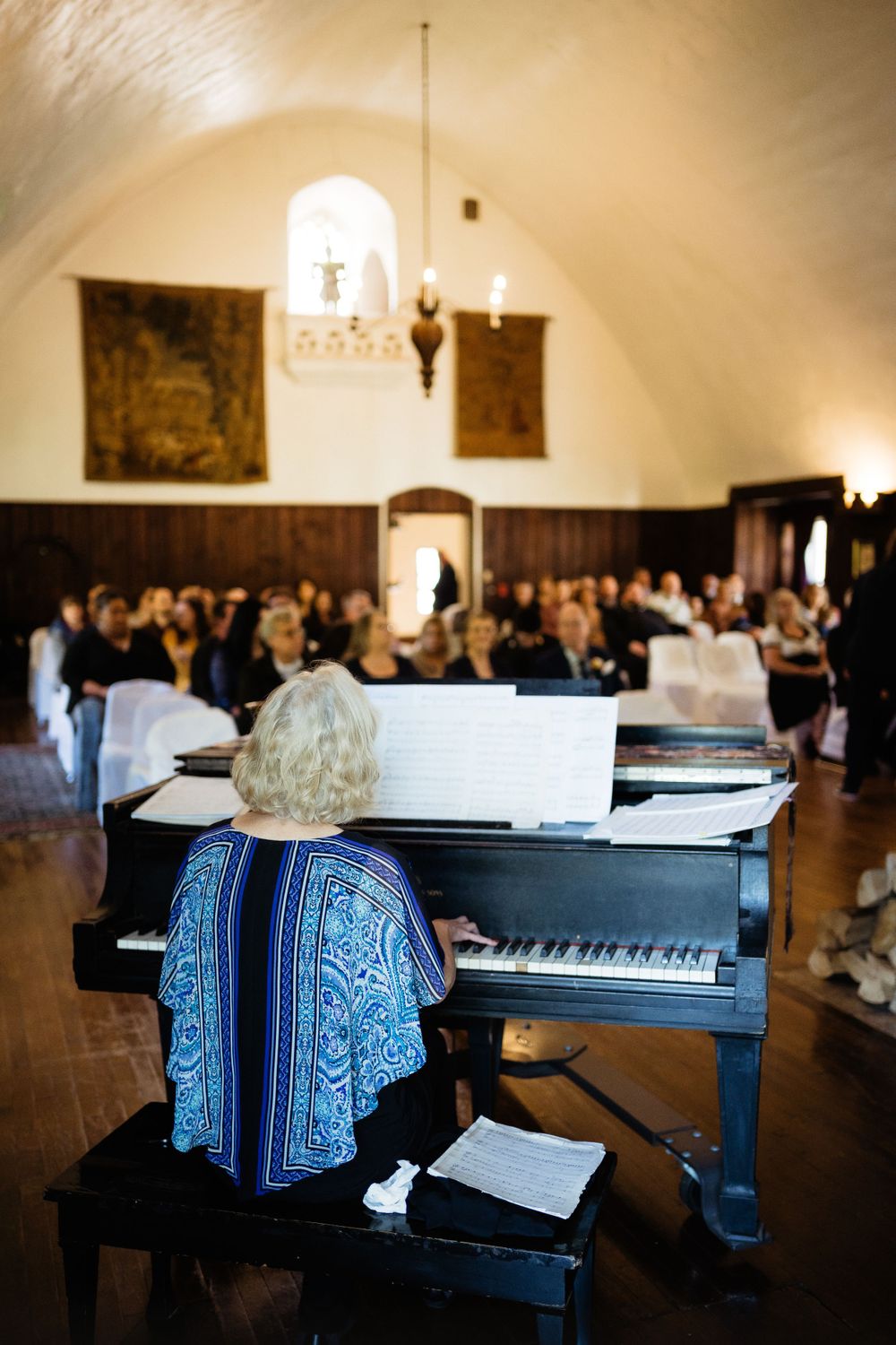 Pianist in a blue shawl performing at a grand piano in an elegant vaulted room.